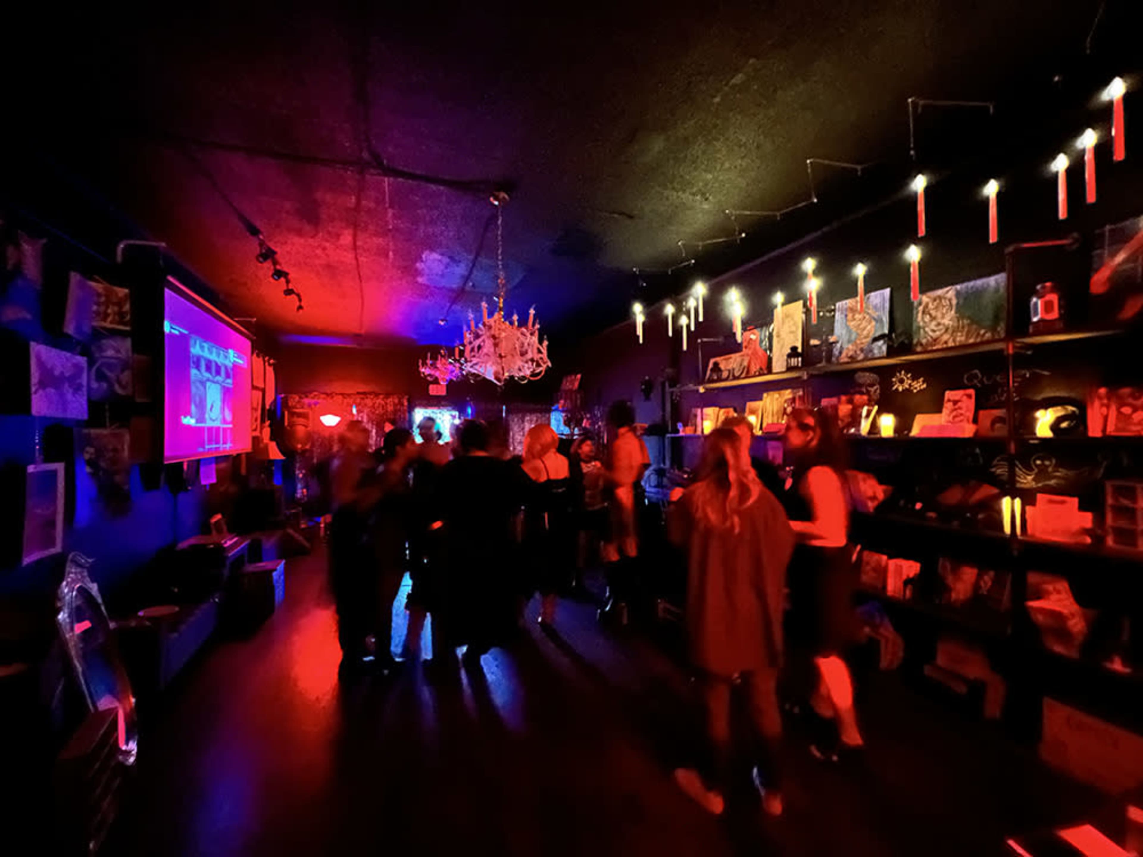 A dimly lit room filled with people socializing, surrounded by colorful artwork and illuminated decor.