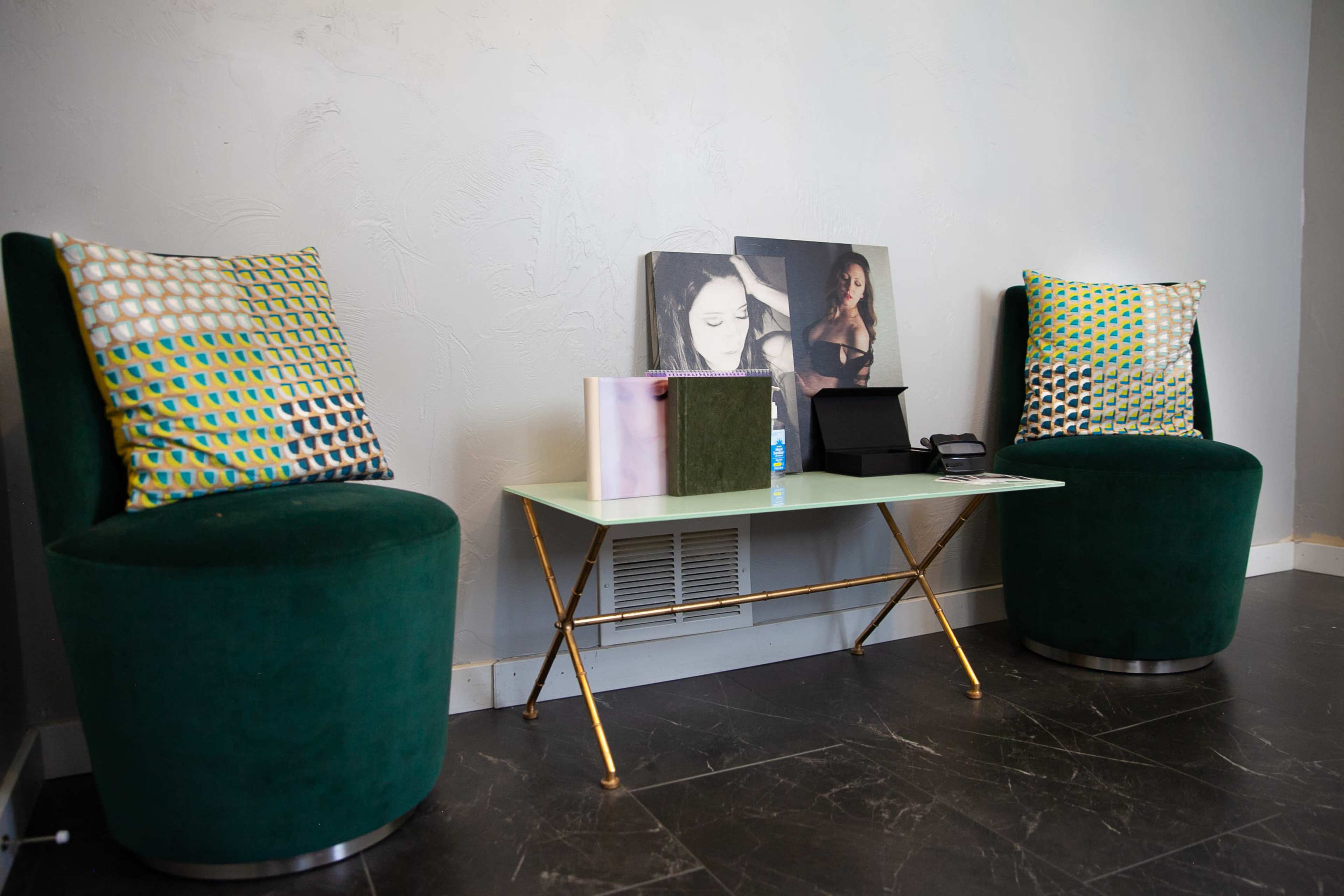 Two green upholstered chairs flank a modern glass table, which holds a black box and a small decorative object, with a wall-mounted artwork featuring a woman in the background.