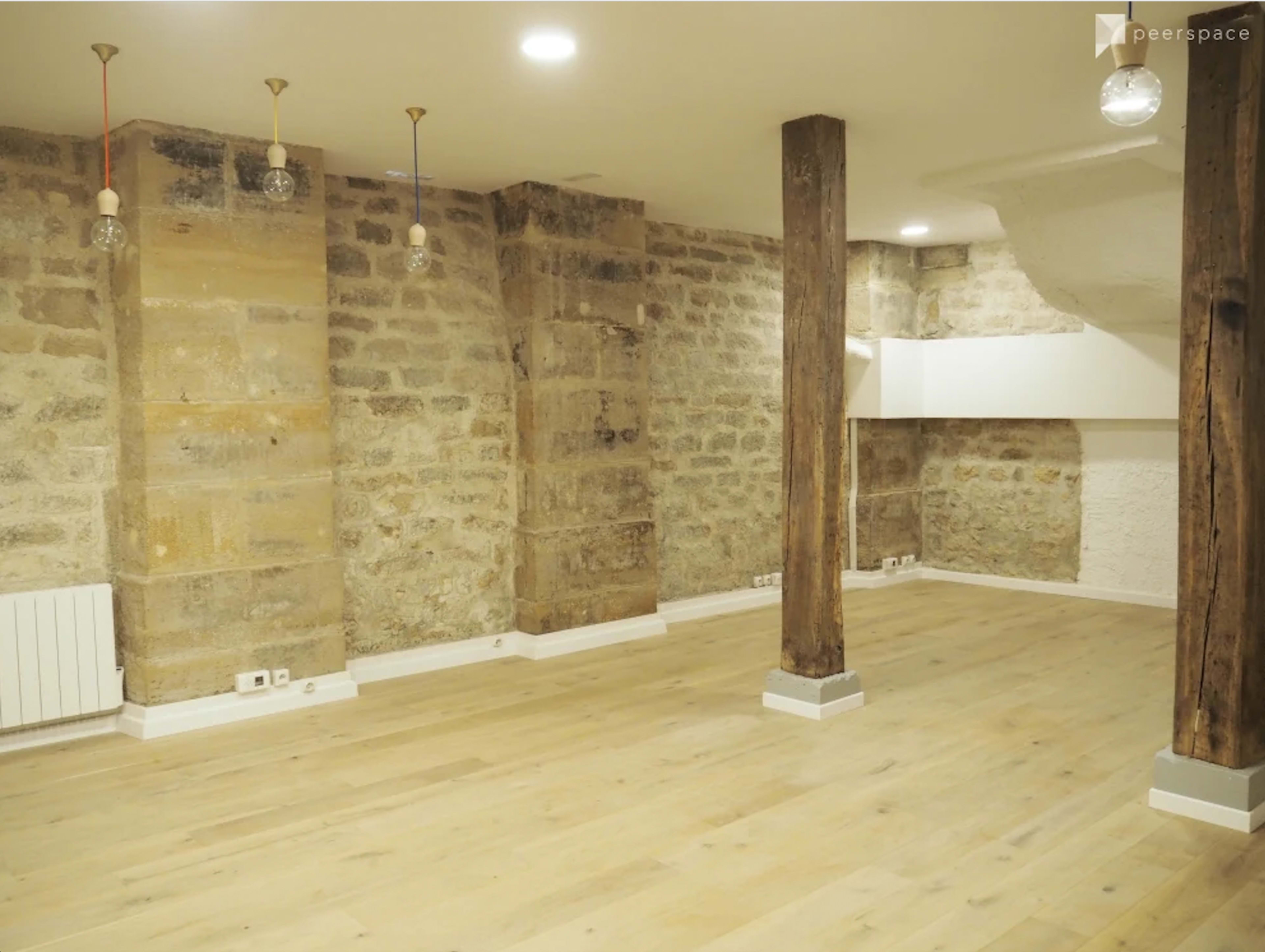 An empty room with stone walls, wooden beams, and wooden flooring, illuminated by pendant lights.