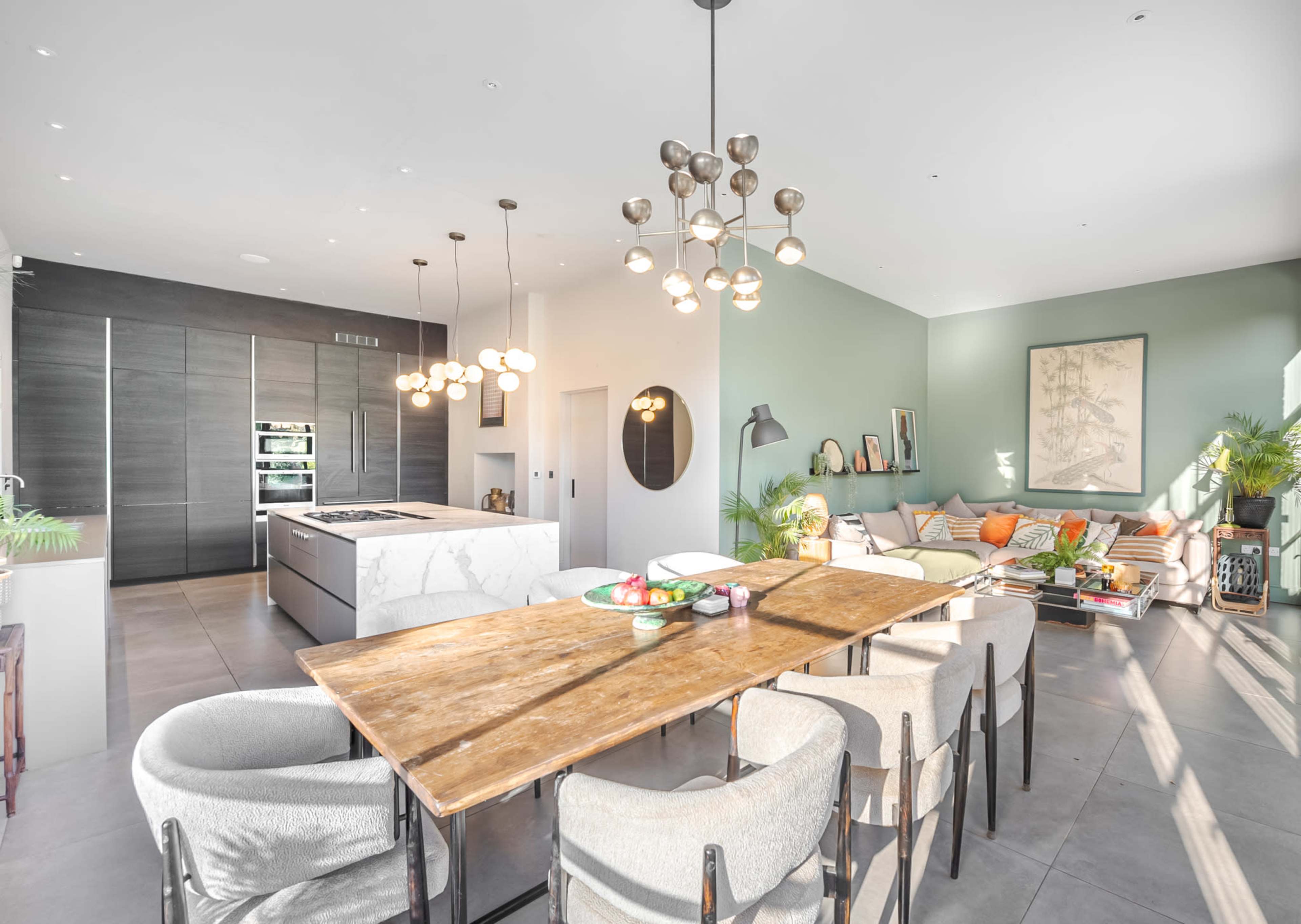 A modern kitchen and dining area featuring a wooden table surrounded by upholstered chairs, a marble kitchen island, and a seating area with a sofa and decorative plants.