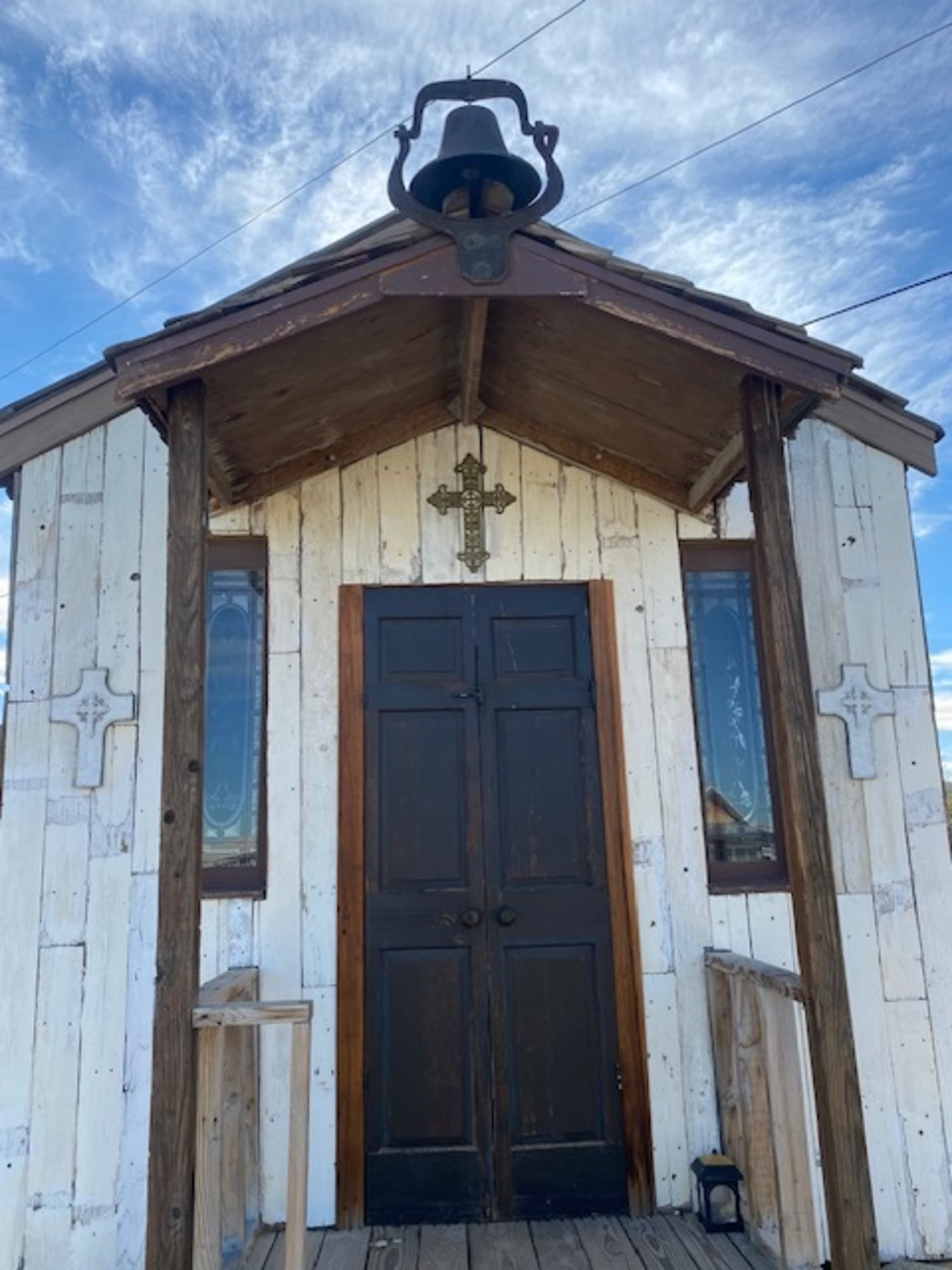 The image shows a small wooden chapel with a dark double door, a bell hanging above the entrance, and decorative crosses on the exterior walls.