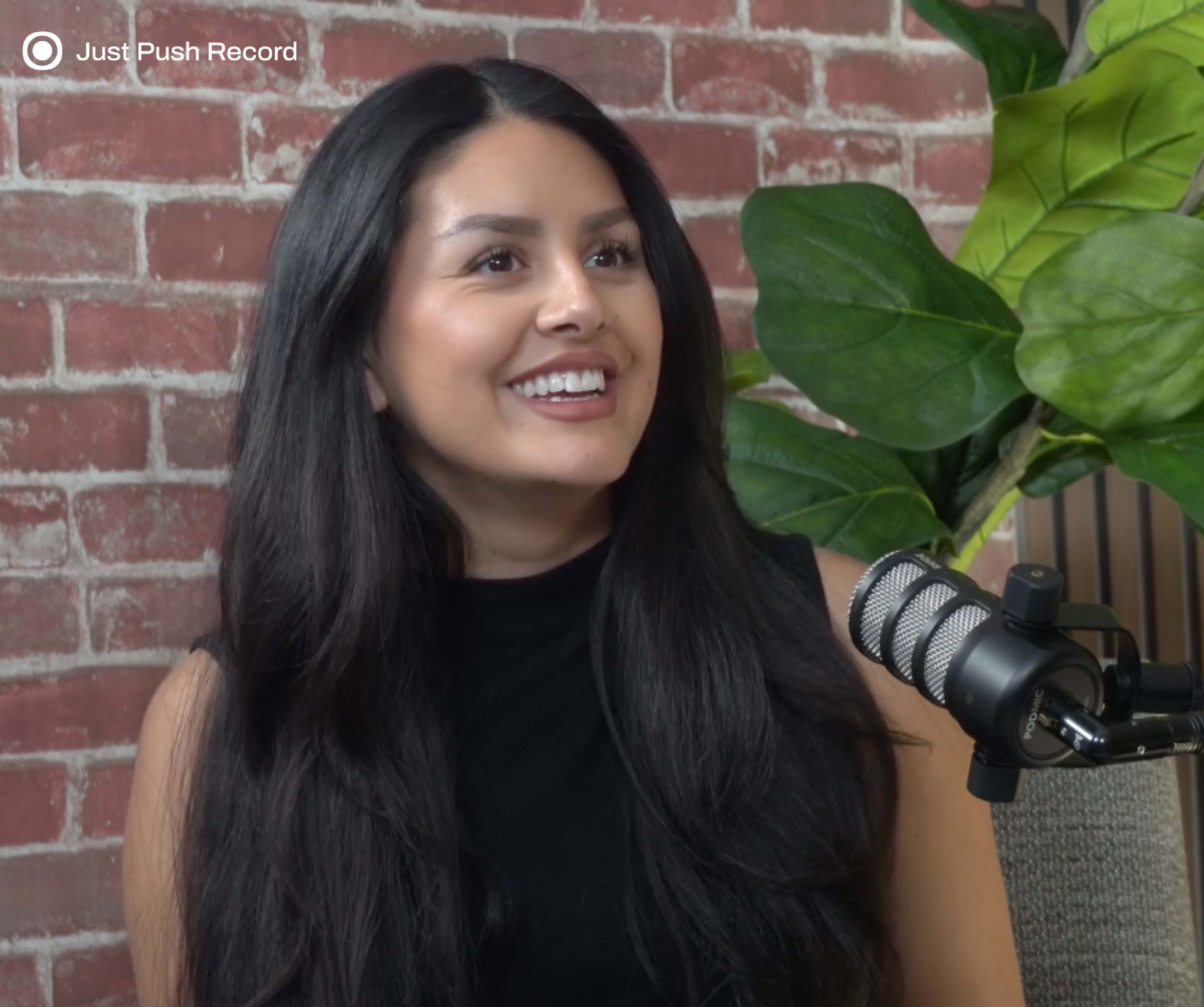 A woman with long black hair is seated in front of a microphone, smiling while being recorded in a room with a brick wall and green plants.
