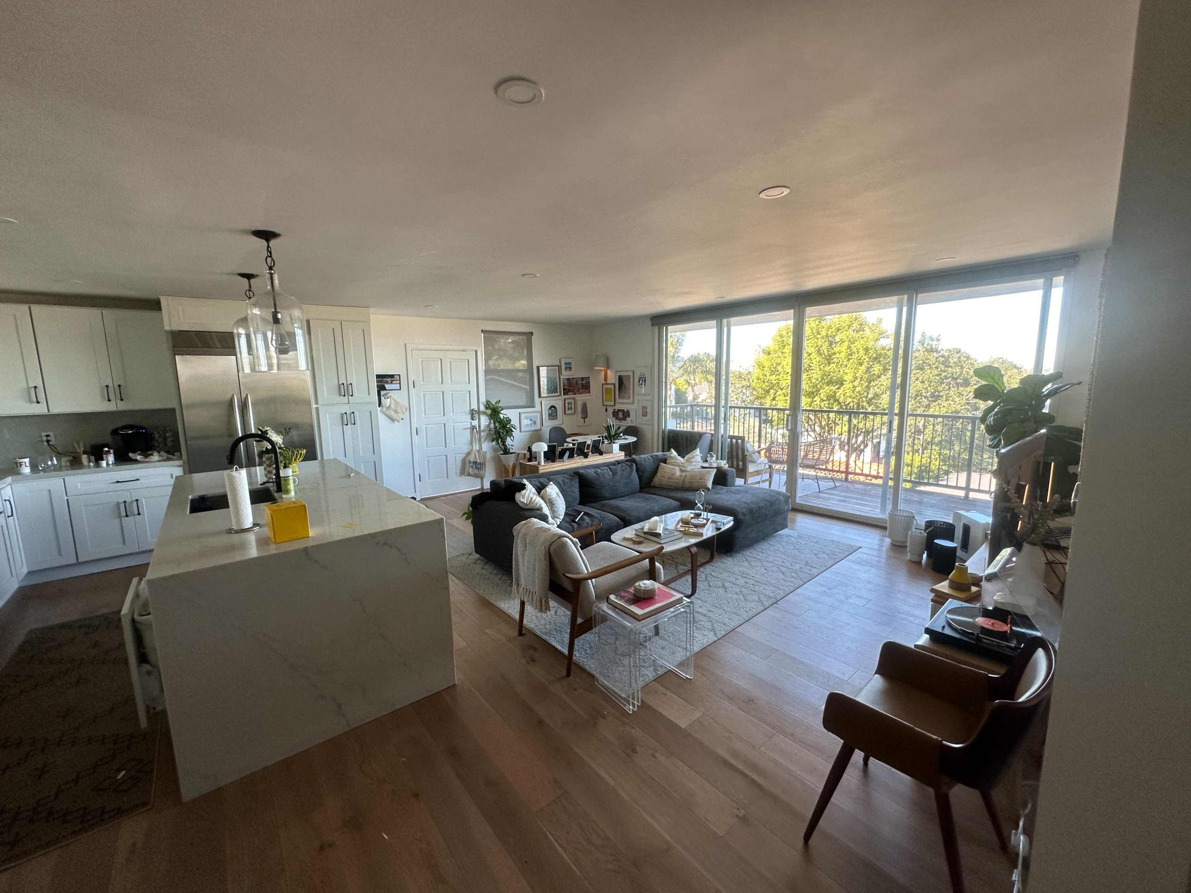 The image shows a modern open-concept living space featuring a kitchen with a large island and a cozy seating area with a sofa and armchairs, all illuminated by natural light from large windows.