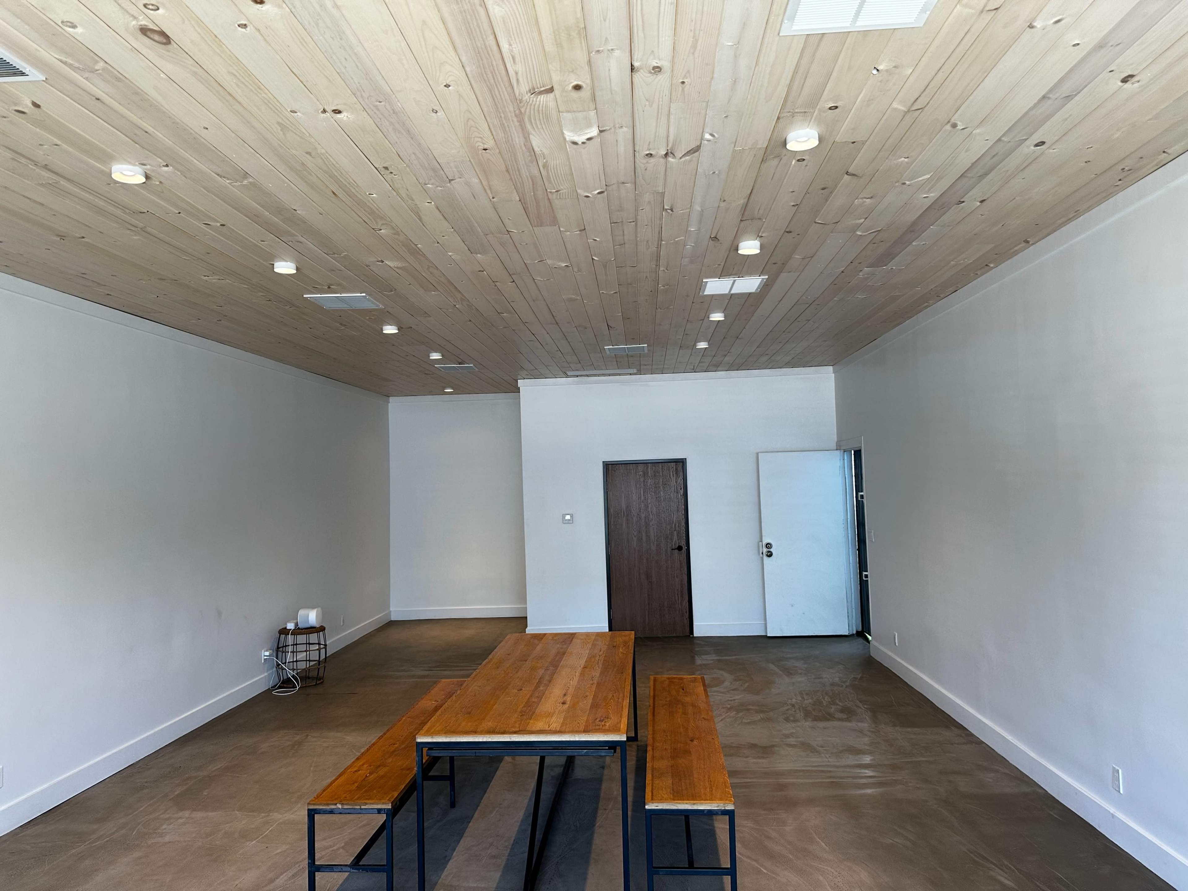 A spacious, minimally furnished room with a wooden table and benches, a plain wall, and a closed door.