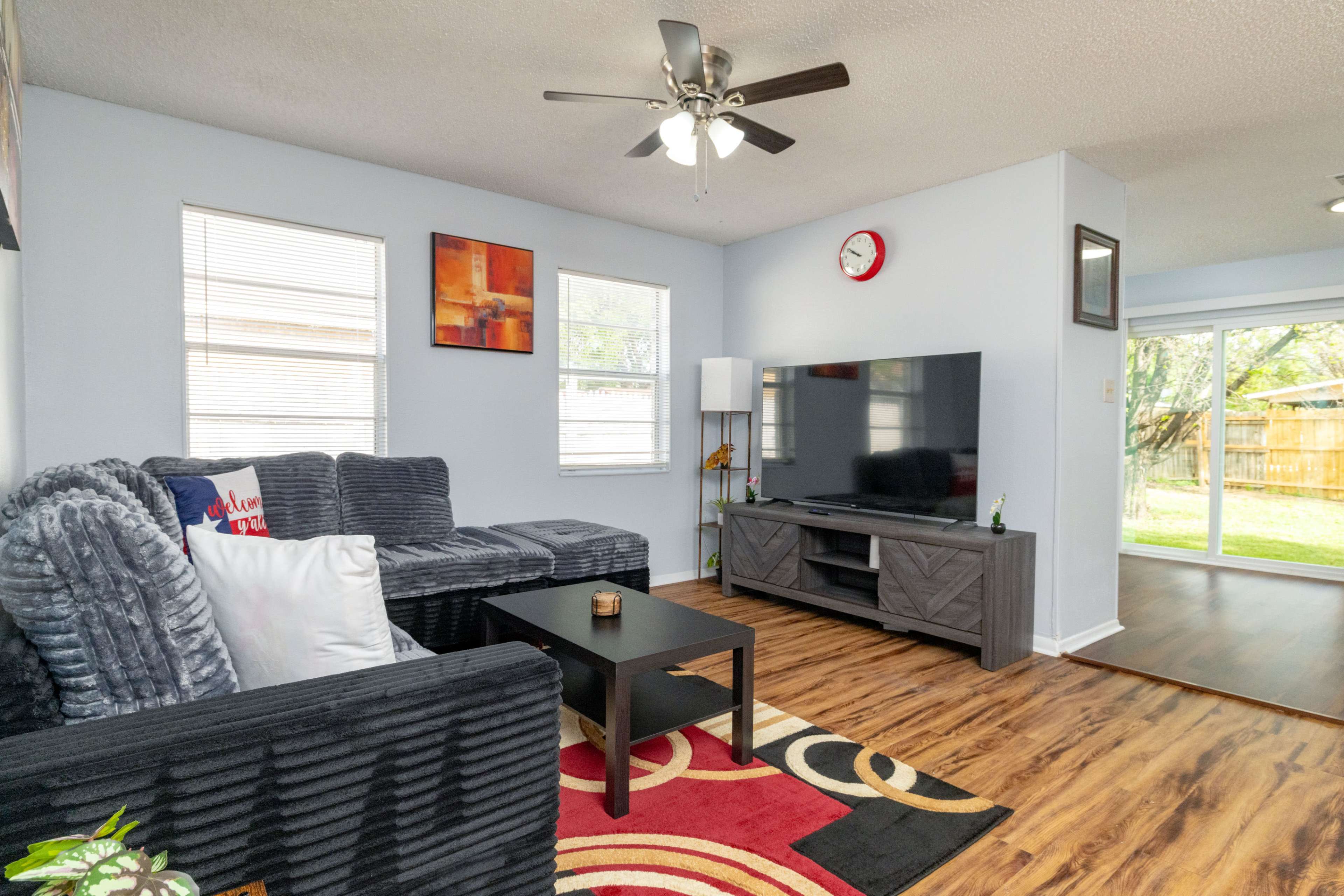 The living room features a sectional sofa, a coffee table, a flat-screen TV, and large windows that provide natural light.