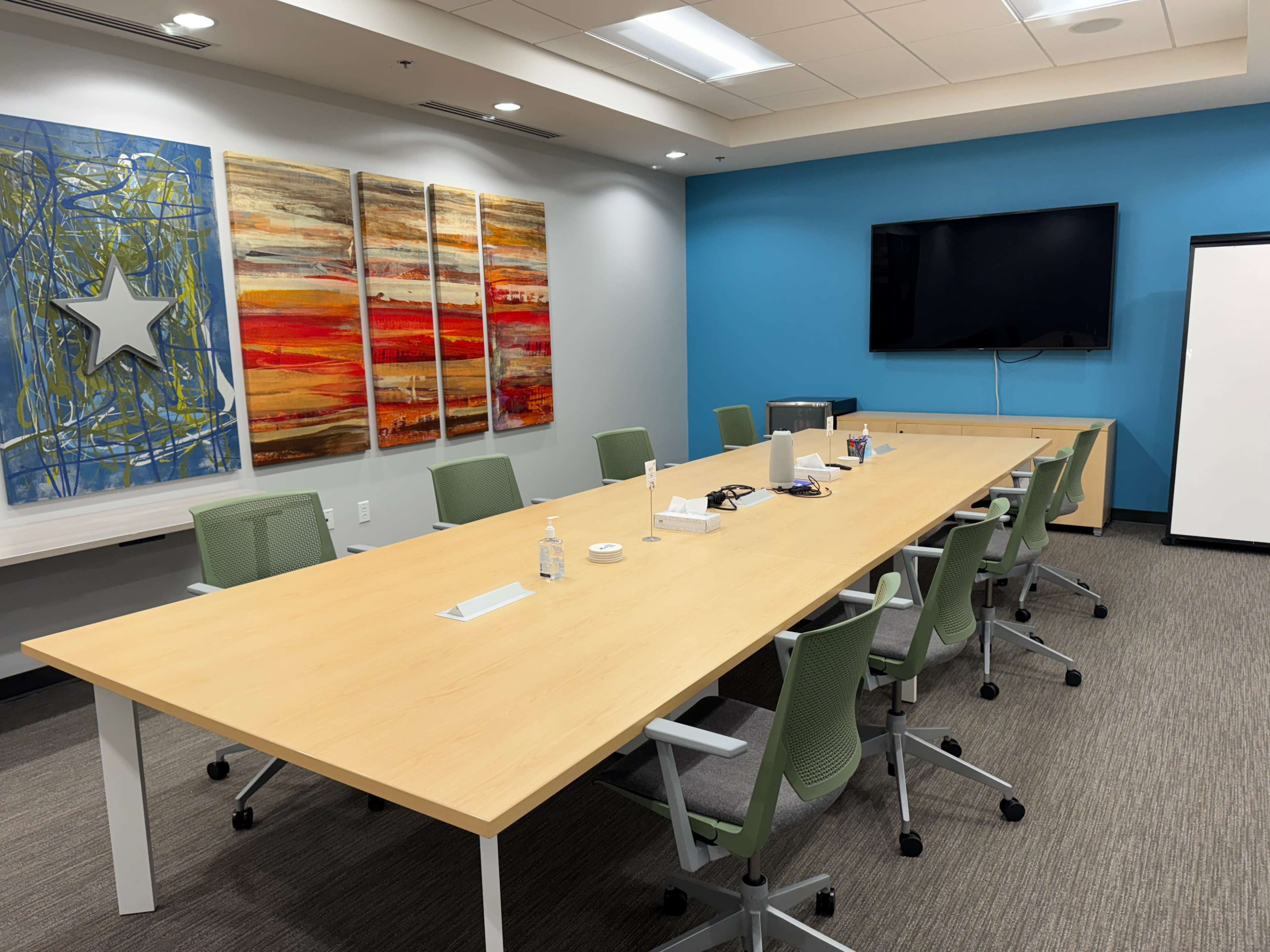 A conference room features a long wooden table surrounded by green chairs, with colorful artwork on the walls and a large screen at one end.