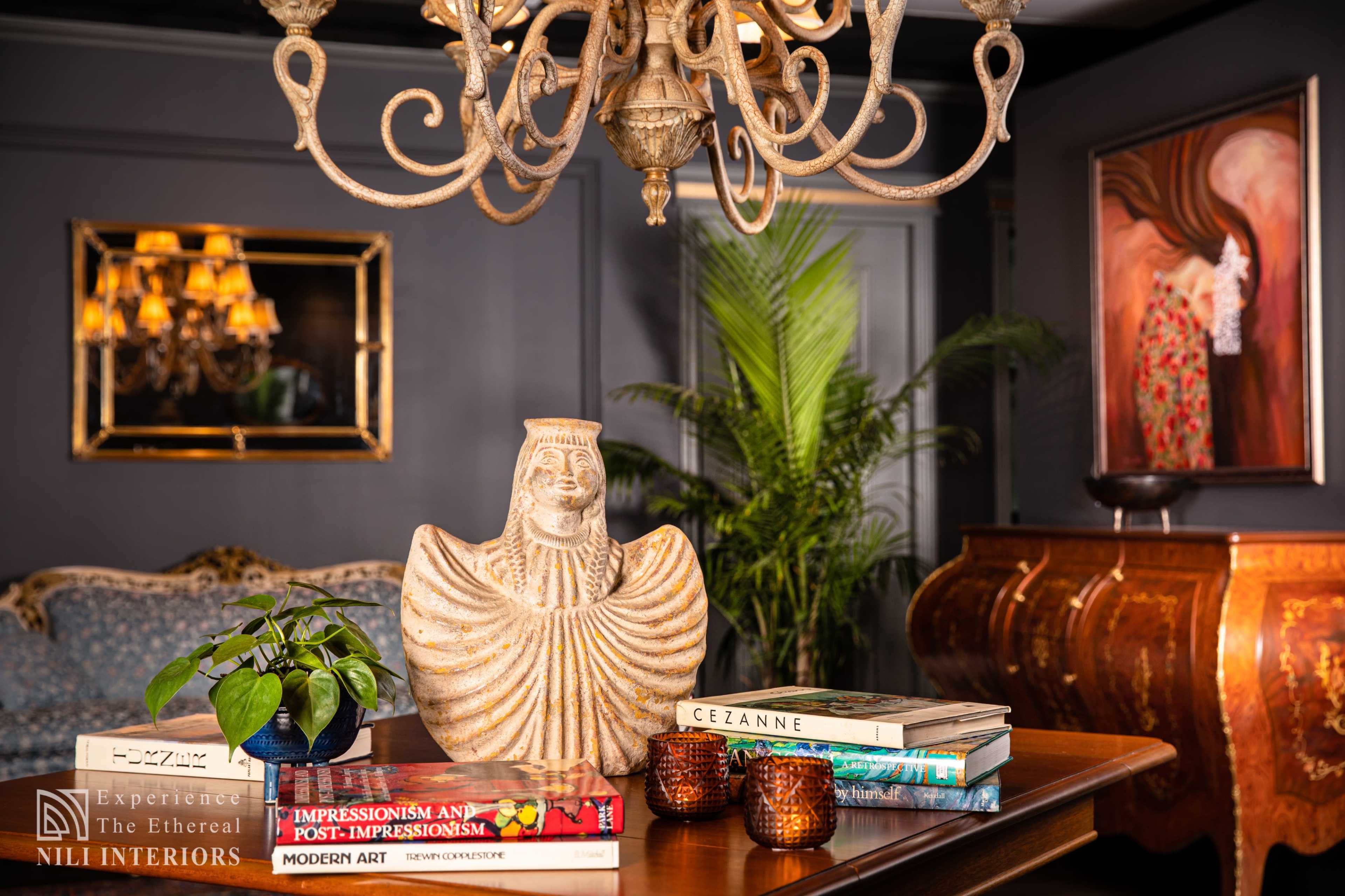 A well-decorated room features a chandelier above a wooden table displaying a decorative vase, books, and a small plant, with upholstered furniture and artwork in the background.