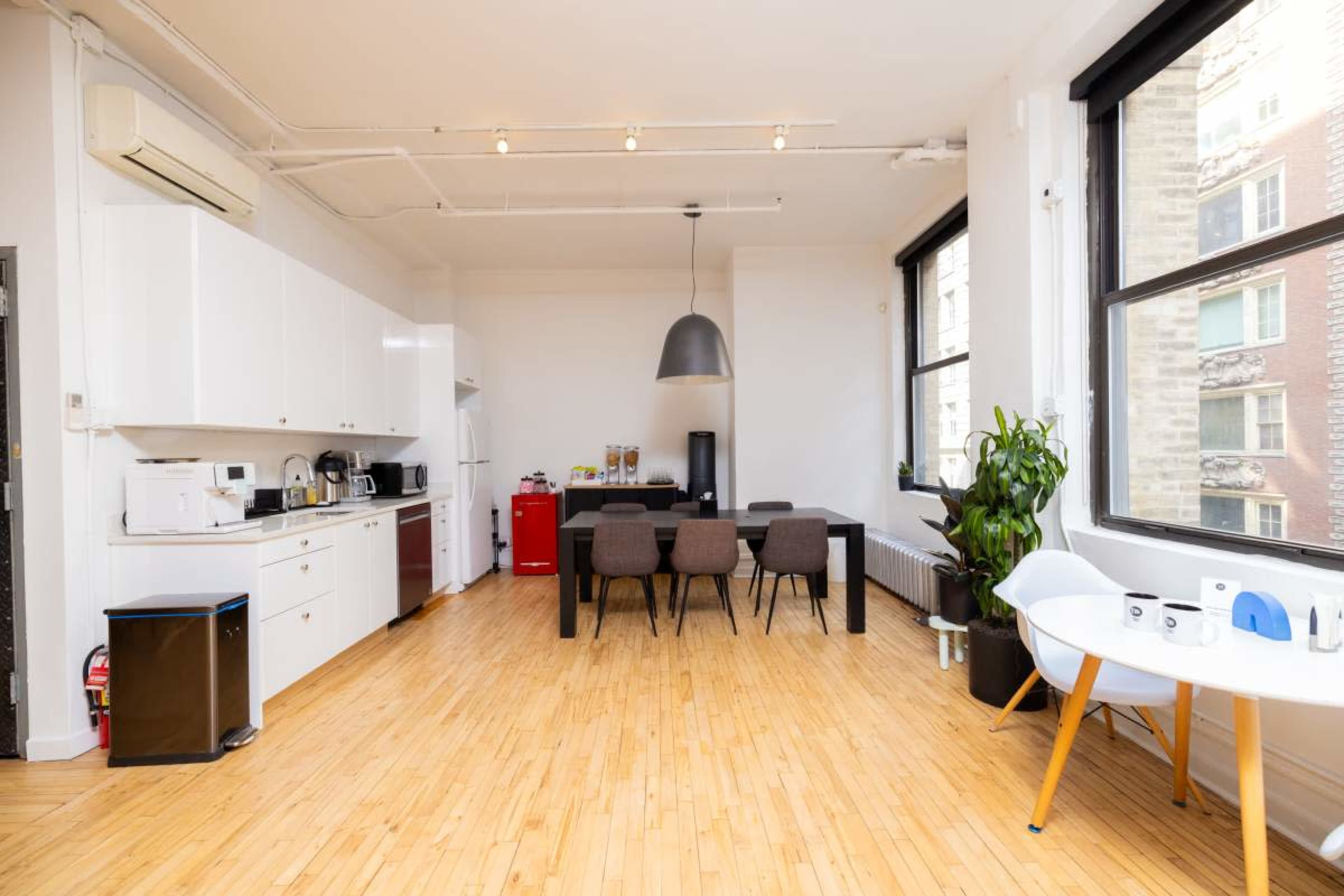 The image shows a bright, open kitchen and dining area with a modern design, featuring a long dining table, a large pendant light, and large windows.