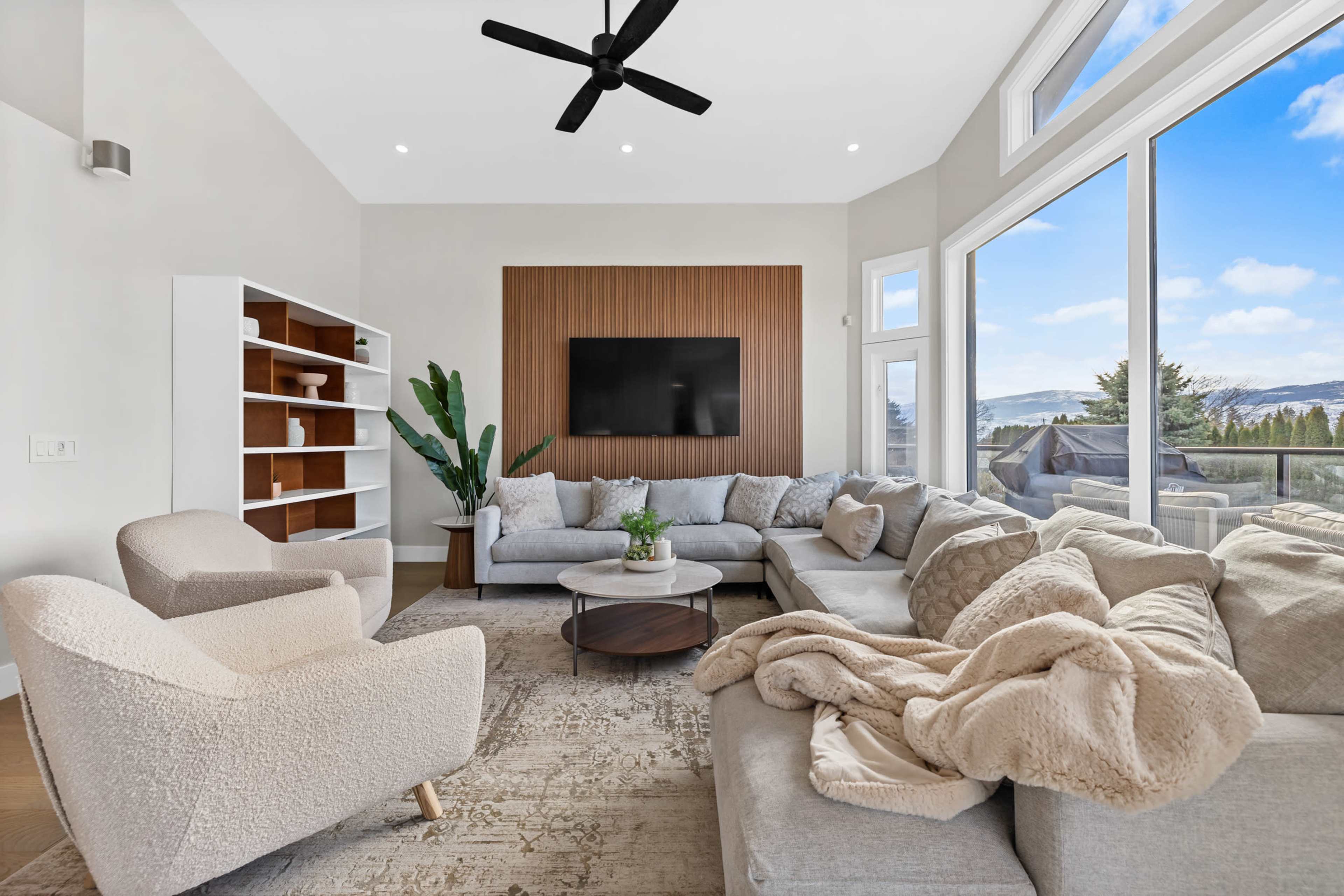 A modern living room with large windows, a sectional sofa, a round coffee table, and a wall-mounted TV, featuring a neutral color palette and minimal decor.