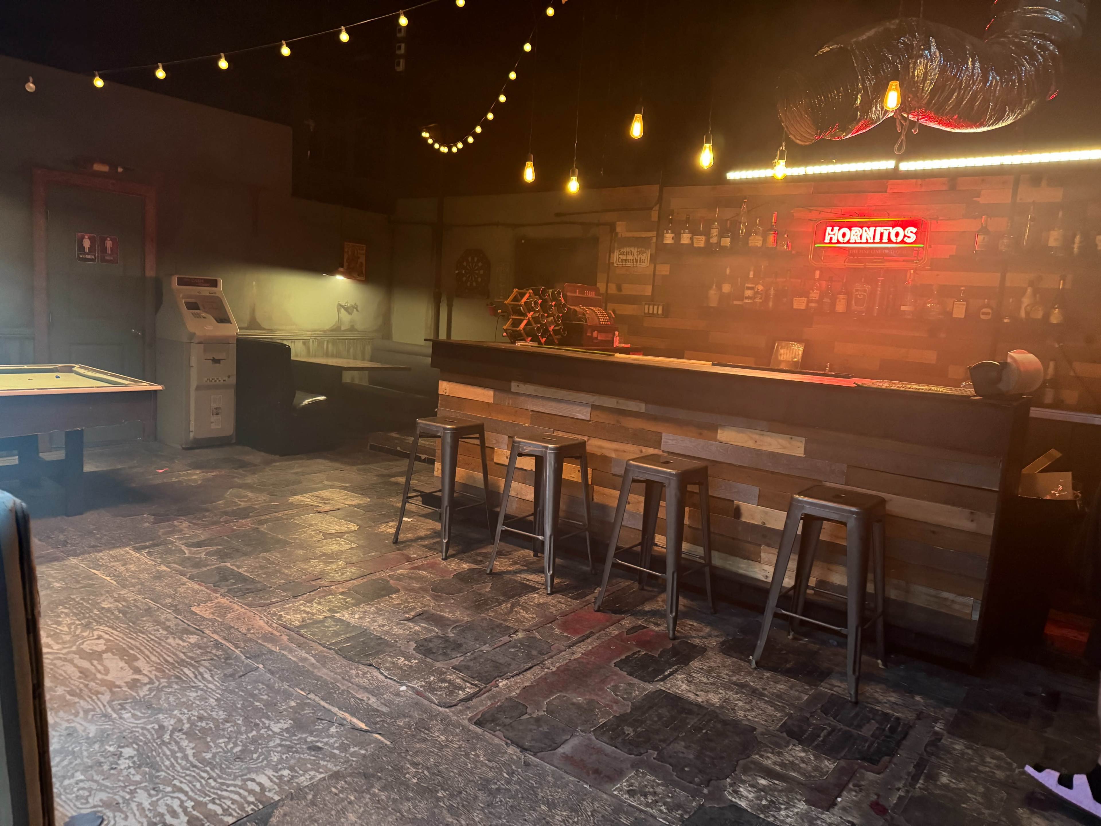 The image shows a dimly lit bar with a wooden counter, a neon sign reading "HORNITOS," and several high stools arranged in front of the bar.