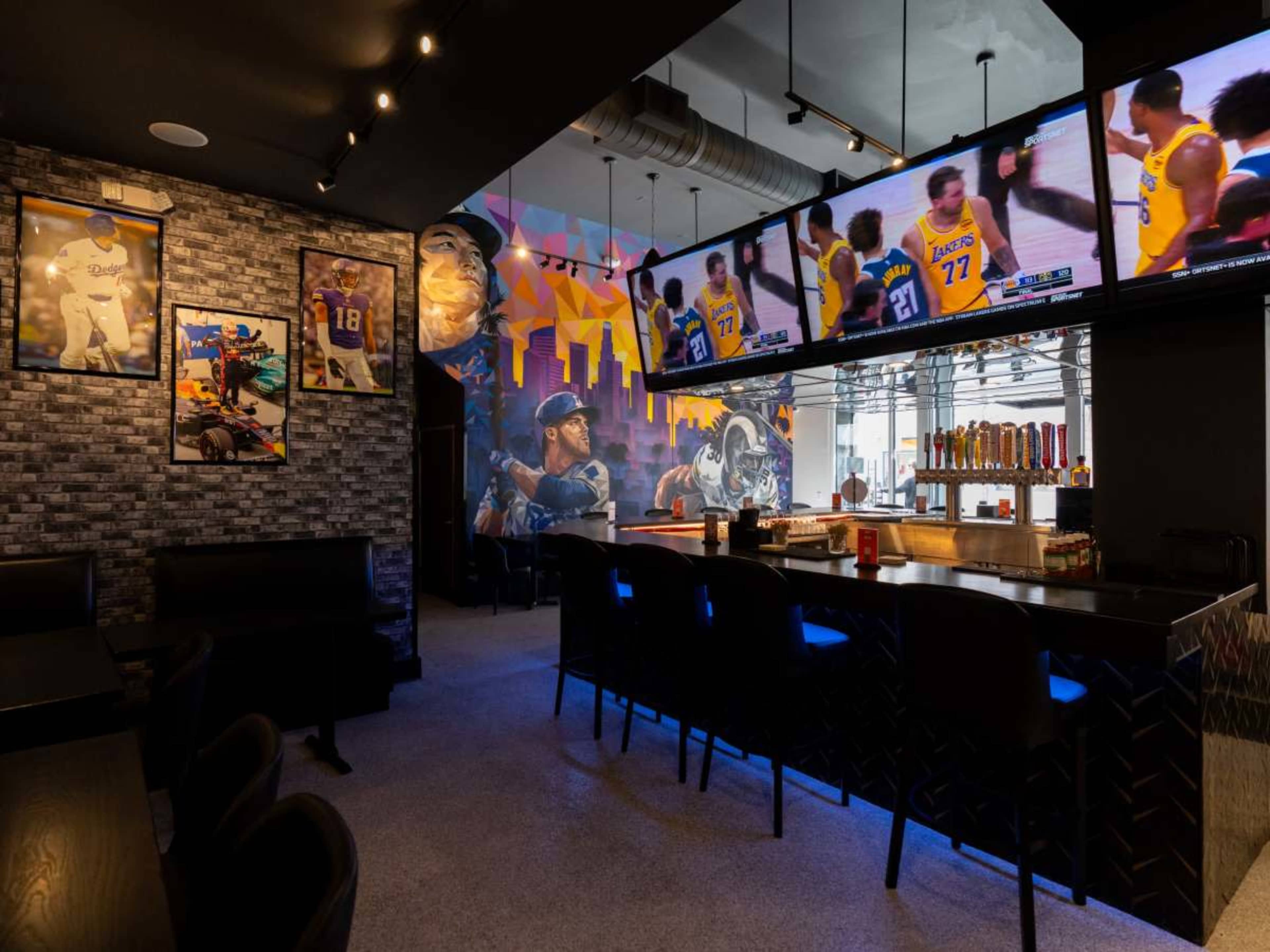 The image shows a modern sports bar featuring multiple television screens displaying a basketball game, murals of athletes on the walls, and a well-stocked bar area with seating.