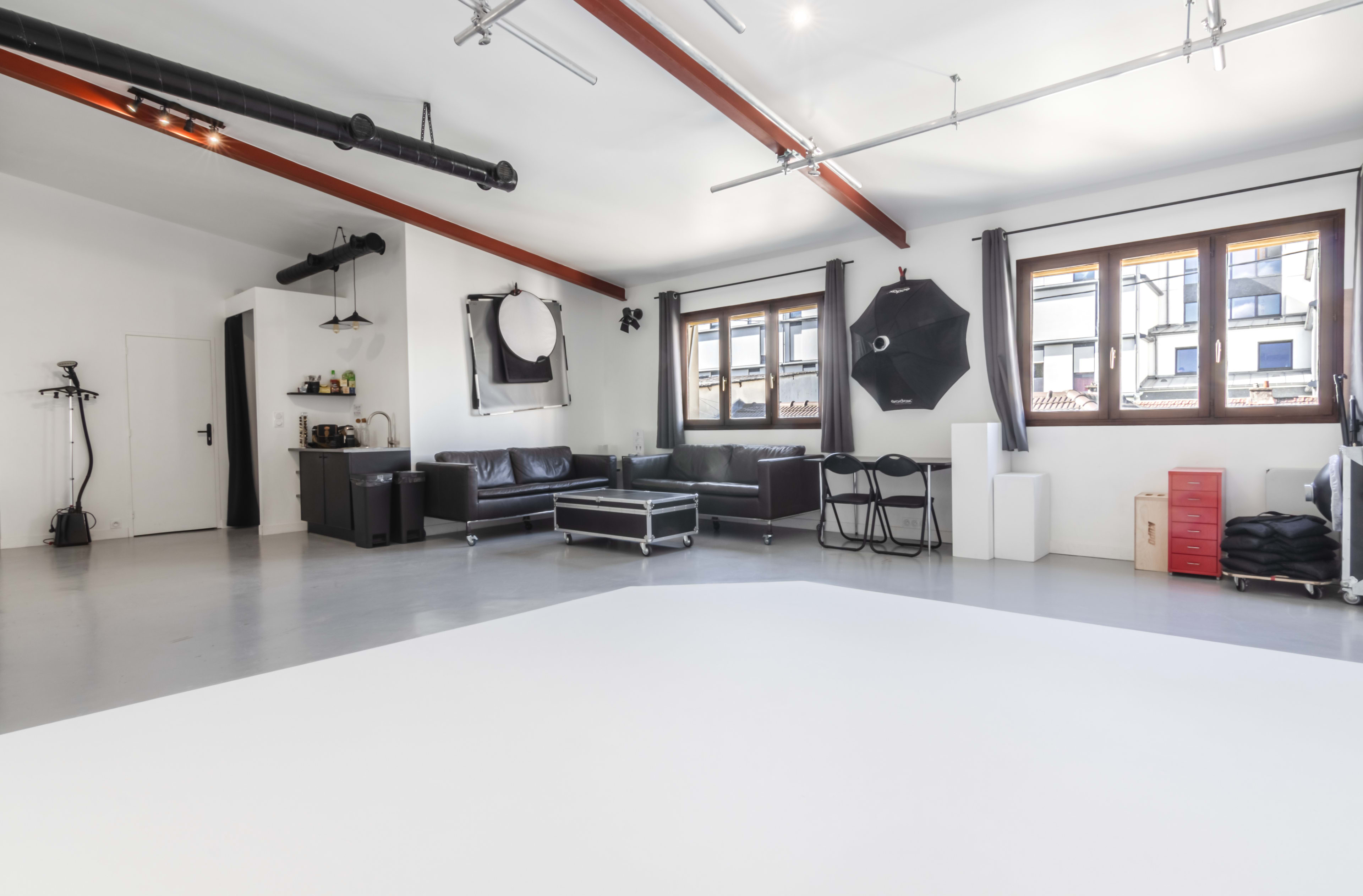 A spacious, modern studio with large windows, a minimalistic black sofa, and photography equipment set up in a well-lit area.