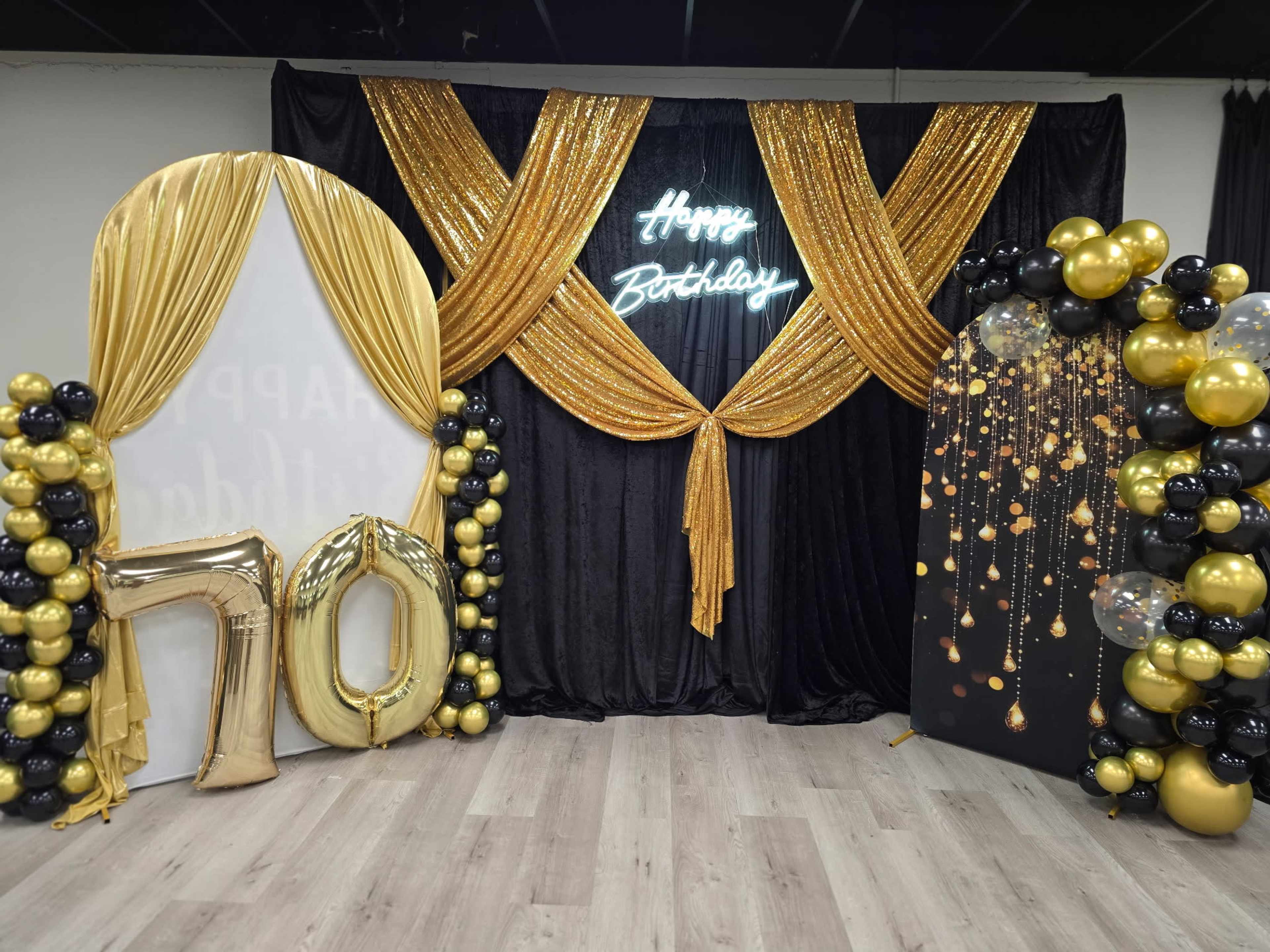 The image features a festive birthday backdrop with gold and black decorations, including a "Happy Birthday" neon sign, a large "70" balloon, and balloon arrangements on either side.