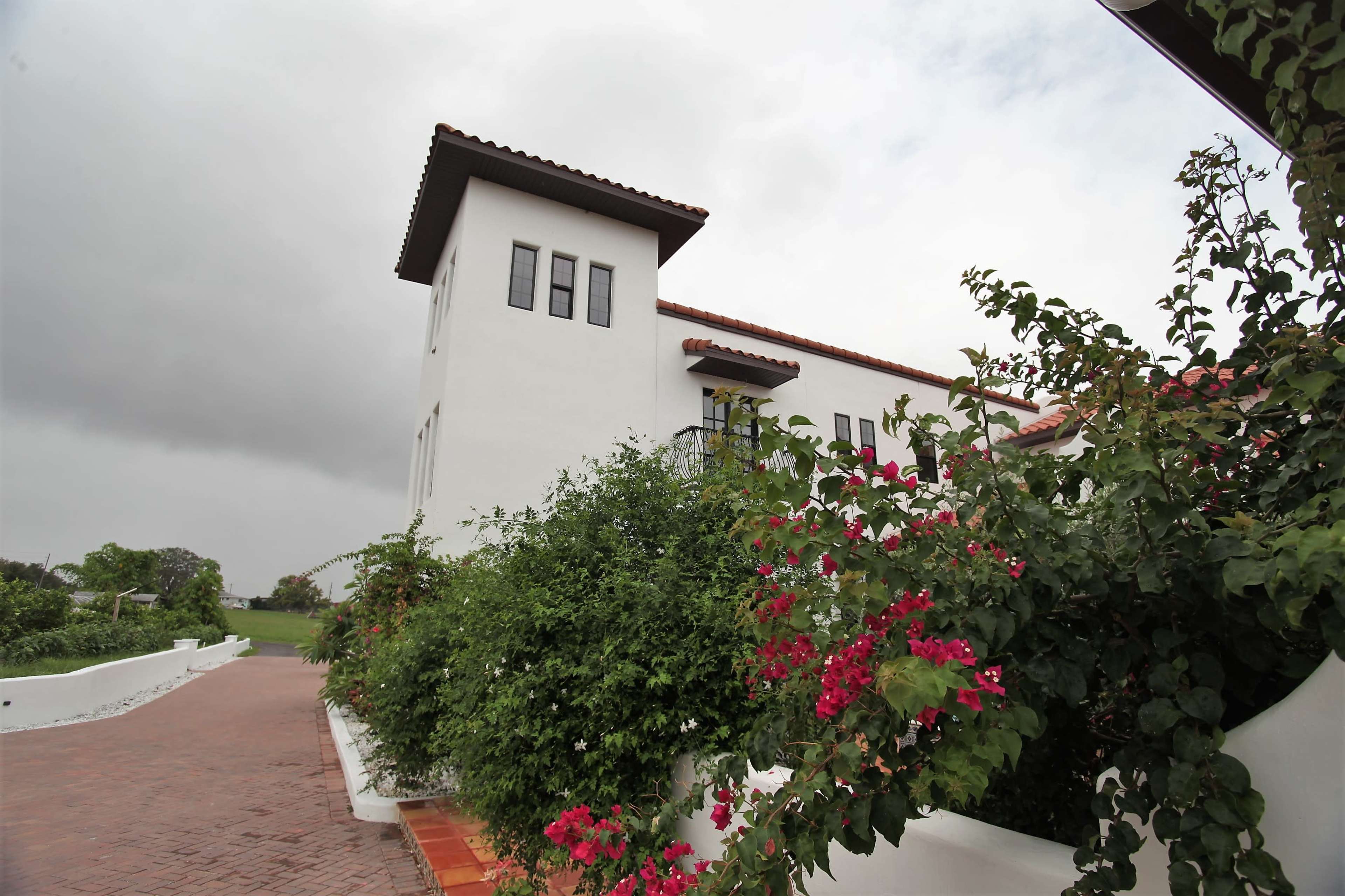 A white stucco building with a tile roof is situated alongside a pathway adorned with vibrant pink bougainvillea.