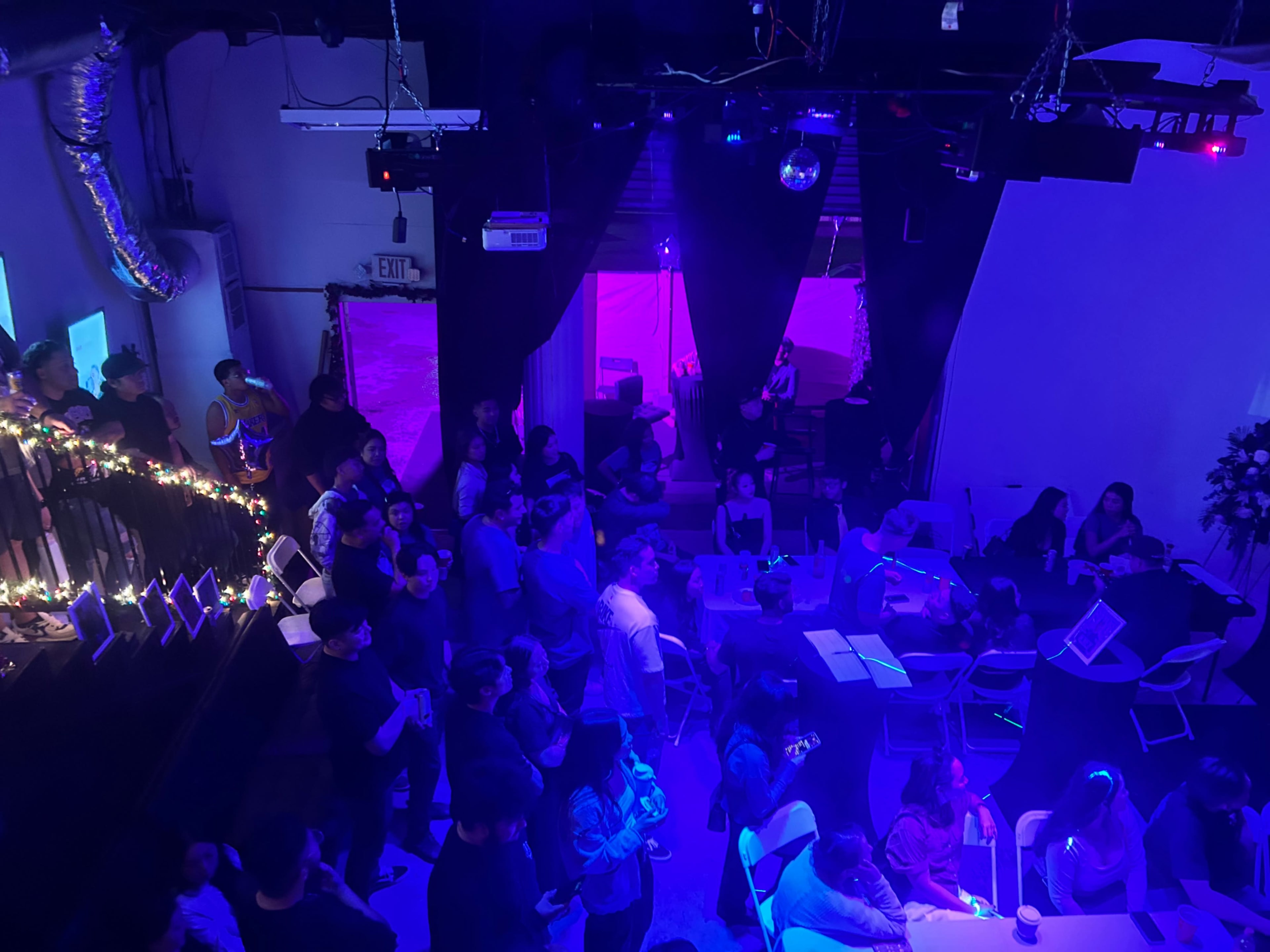 A crowd of people is gathered in a dimly lit space with purple and blue lighting, while some are seated at tables and others stand near the walls.