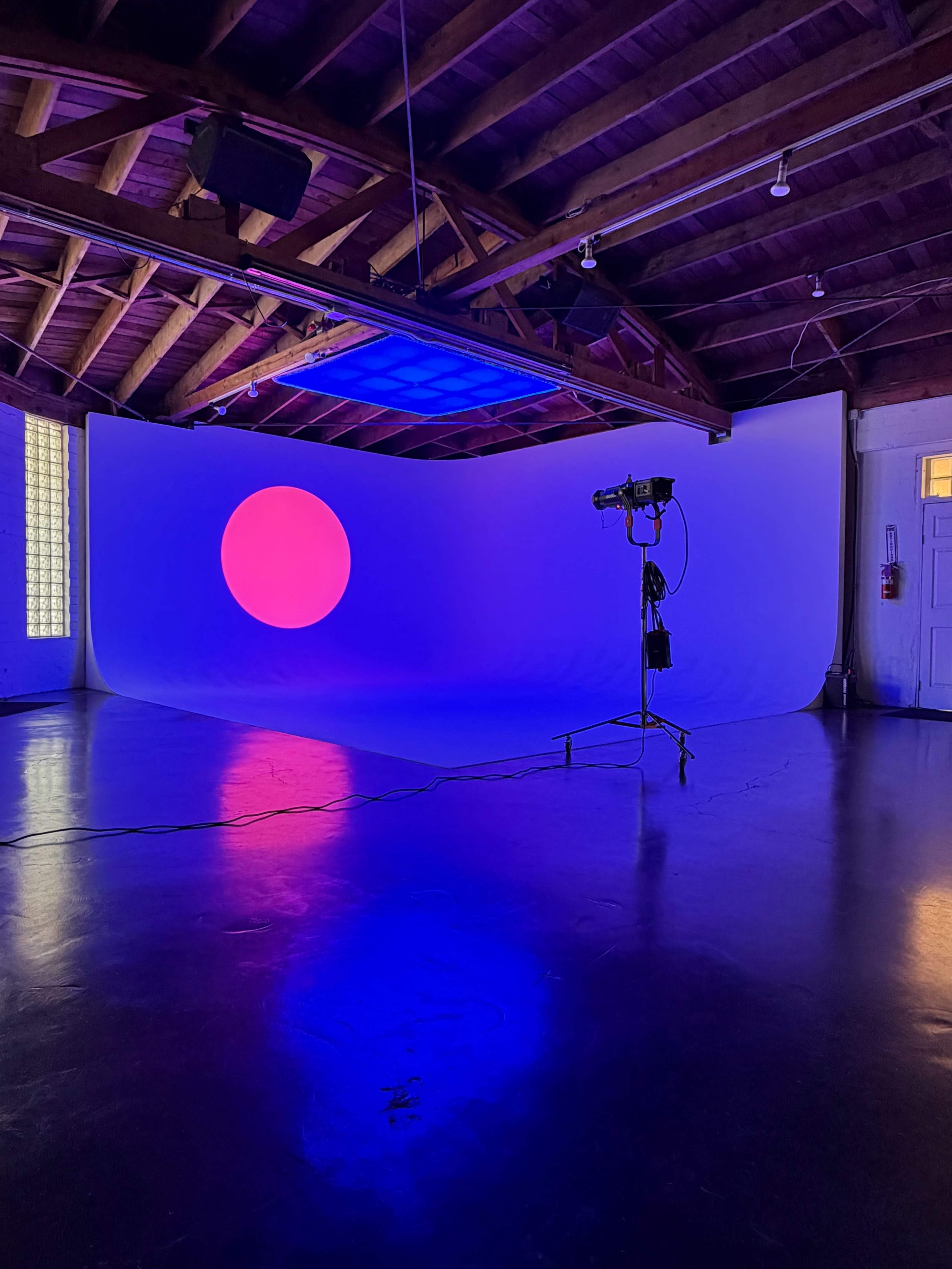The image shows an empty studio space with a large curved backdrop illuminated by a purple circle and soft lighting, featuring wooden beams on the ceiling.