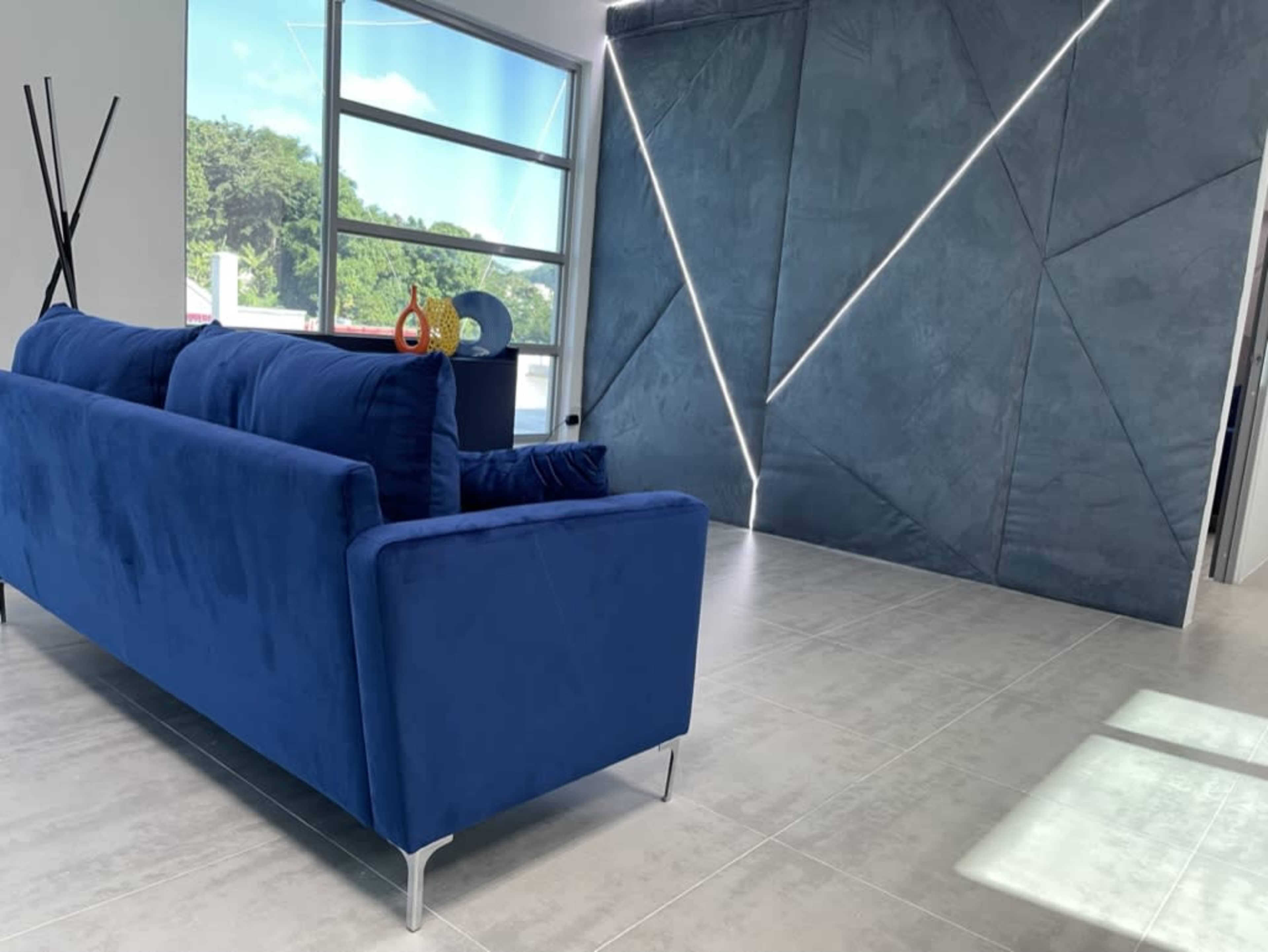 A blue couch is positioned in a modern room with large windows and a geometric wall design illuminated by thin lights.