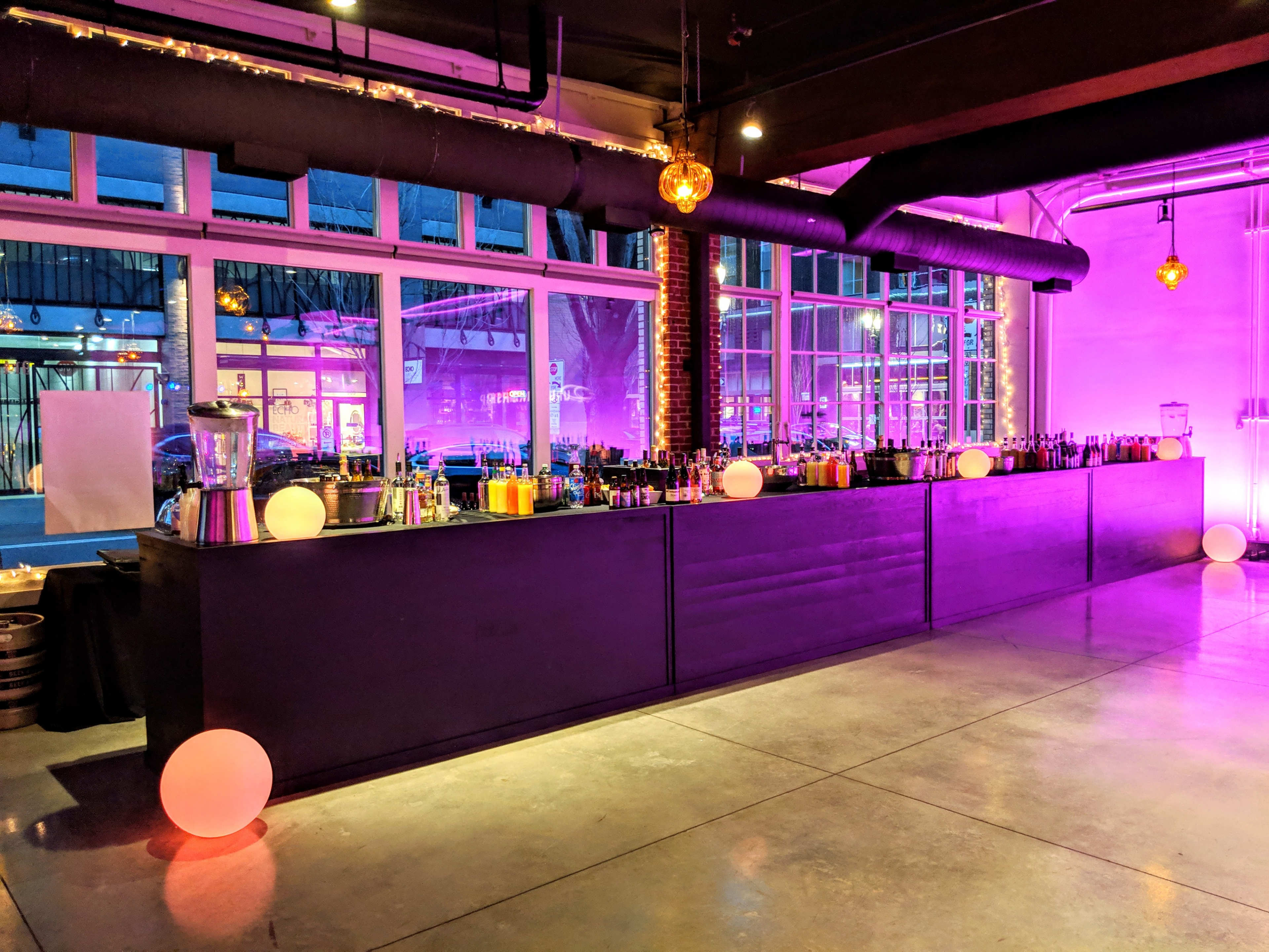 A modern bar area with a long counter filled with various bottles and illuminated by purple lighting, alongside round light fixtures on the floor.