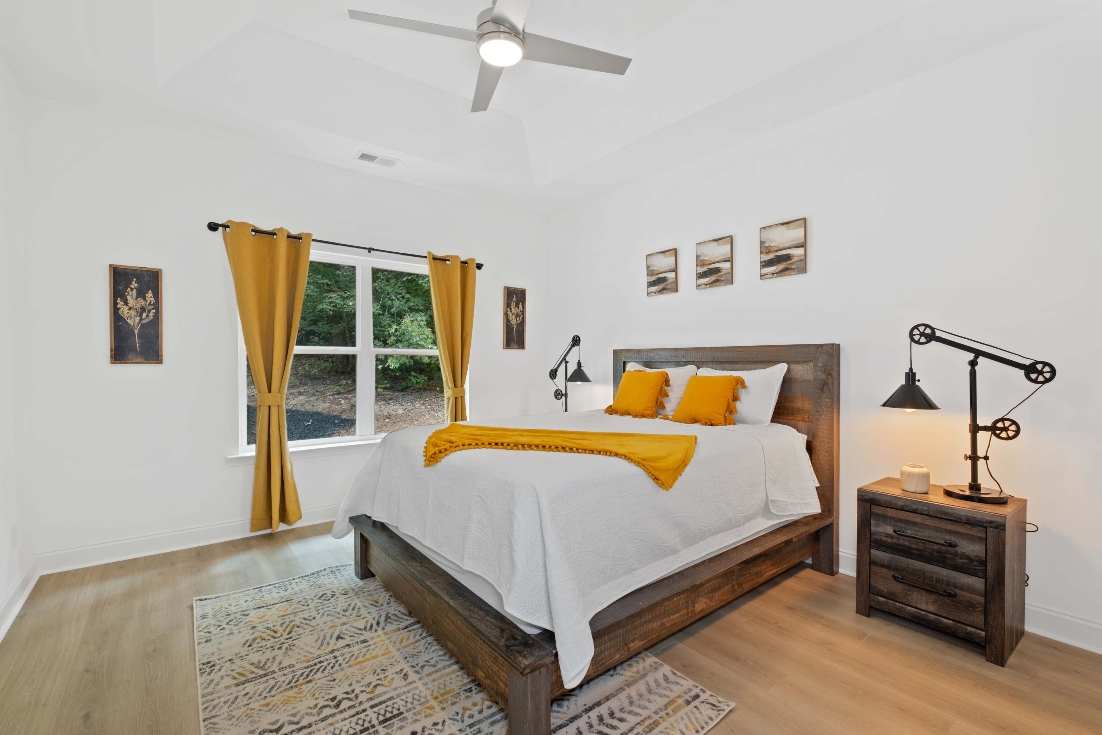 A bright bedroom featuring a wooden bed with white bedding, yellow pillows, and two side tables, accompanied by large windows dressed with yellow curtains.