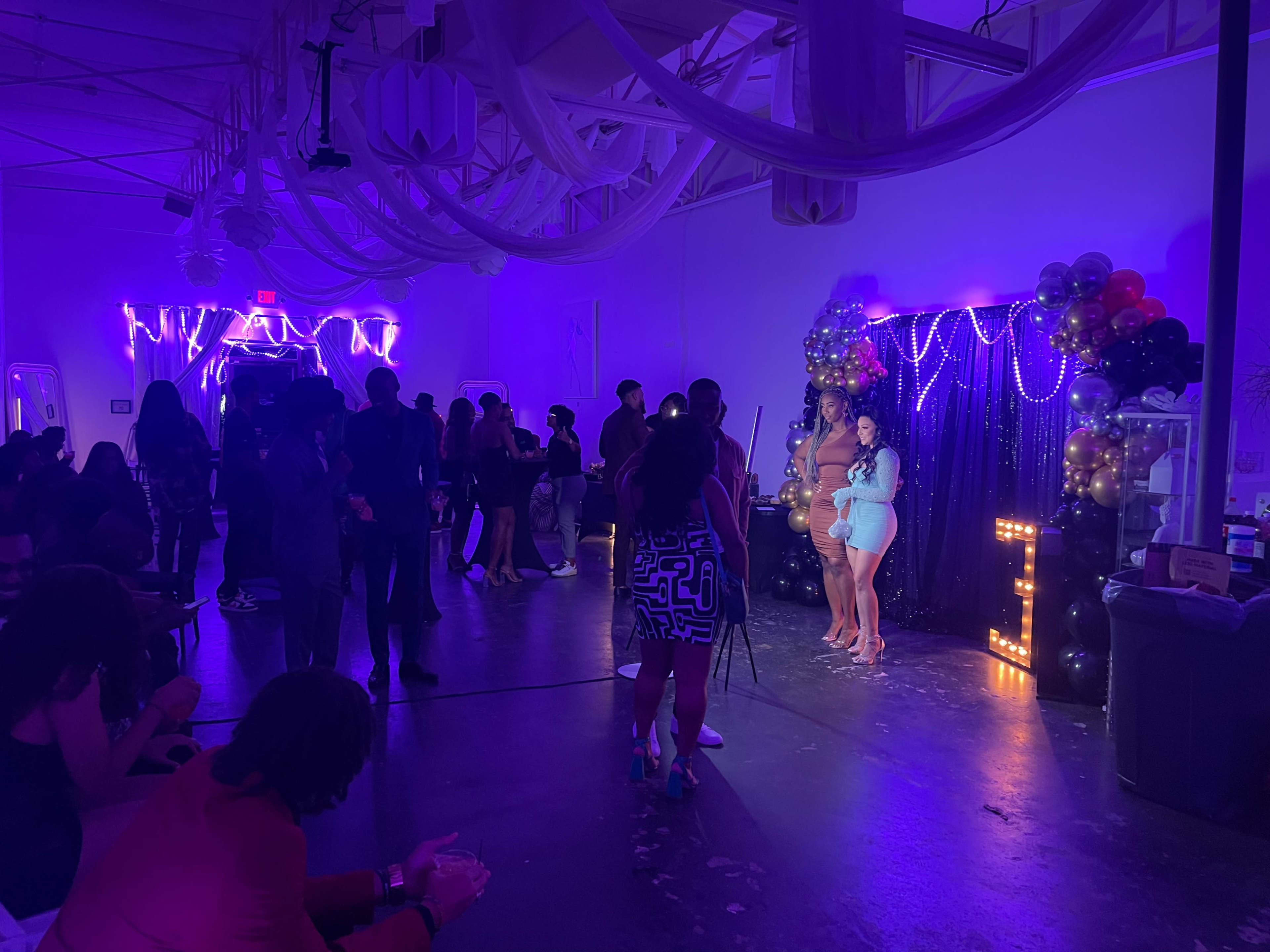 The image shows a vibrant party scene with guests mingling under purple lighting, while a group poses for a photo in front of a decorative backdrop adorned with balloons and lights.