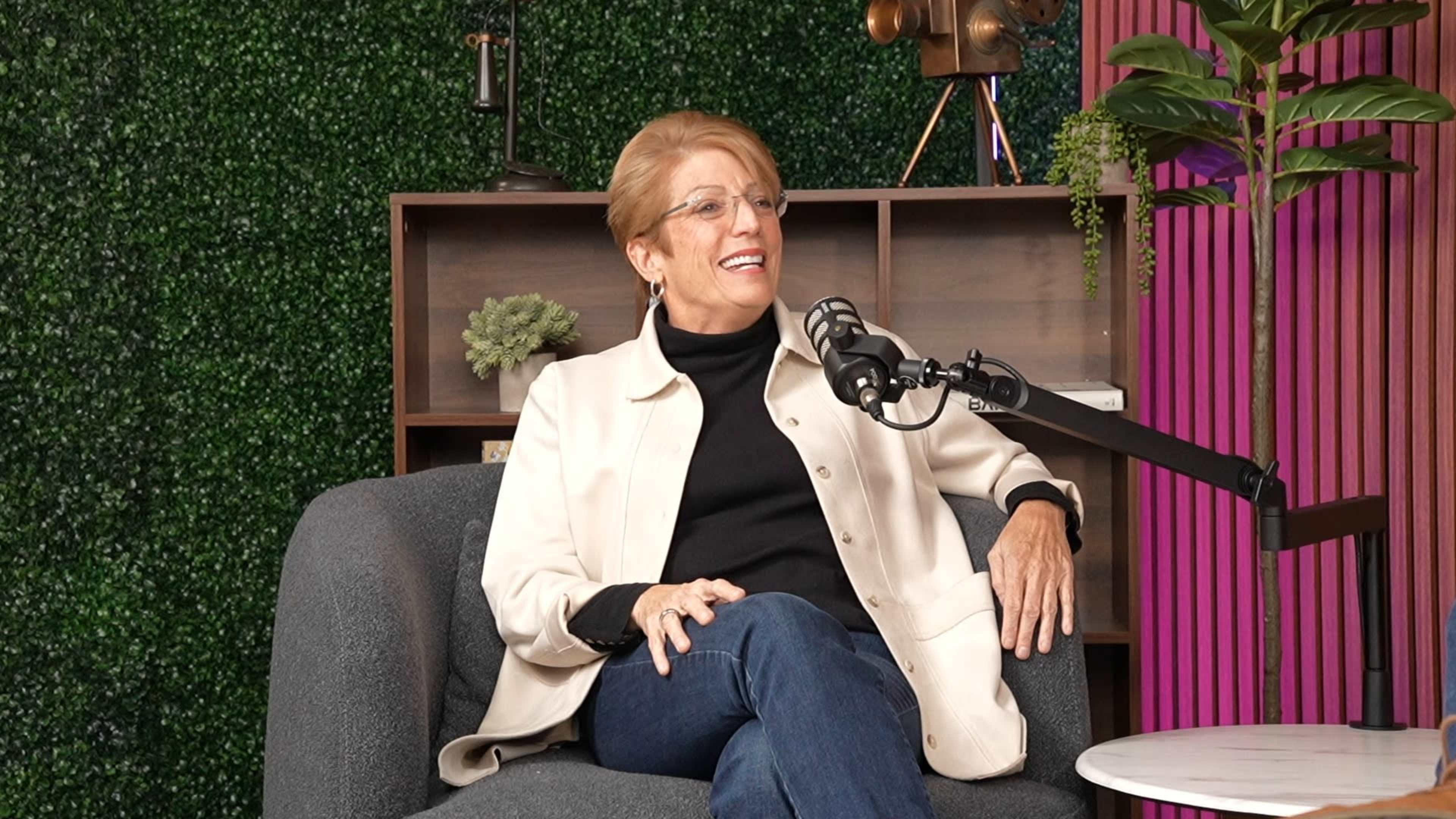 A woman in a light-colored jacket sits on a gray couch in a studio setting, smiling and engaged in conversation near a microphone.