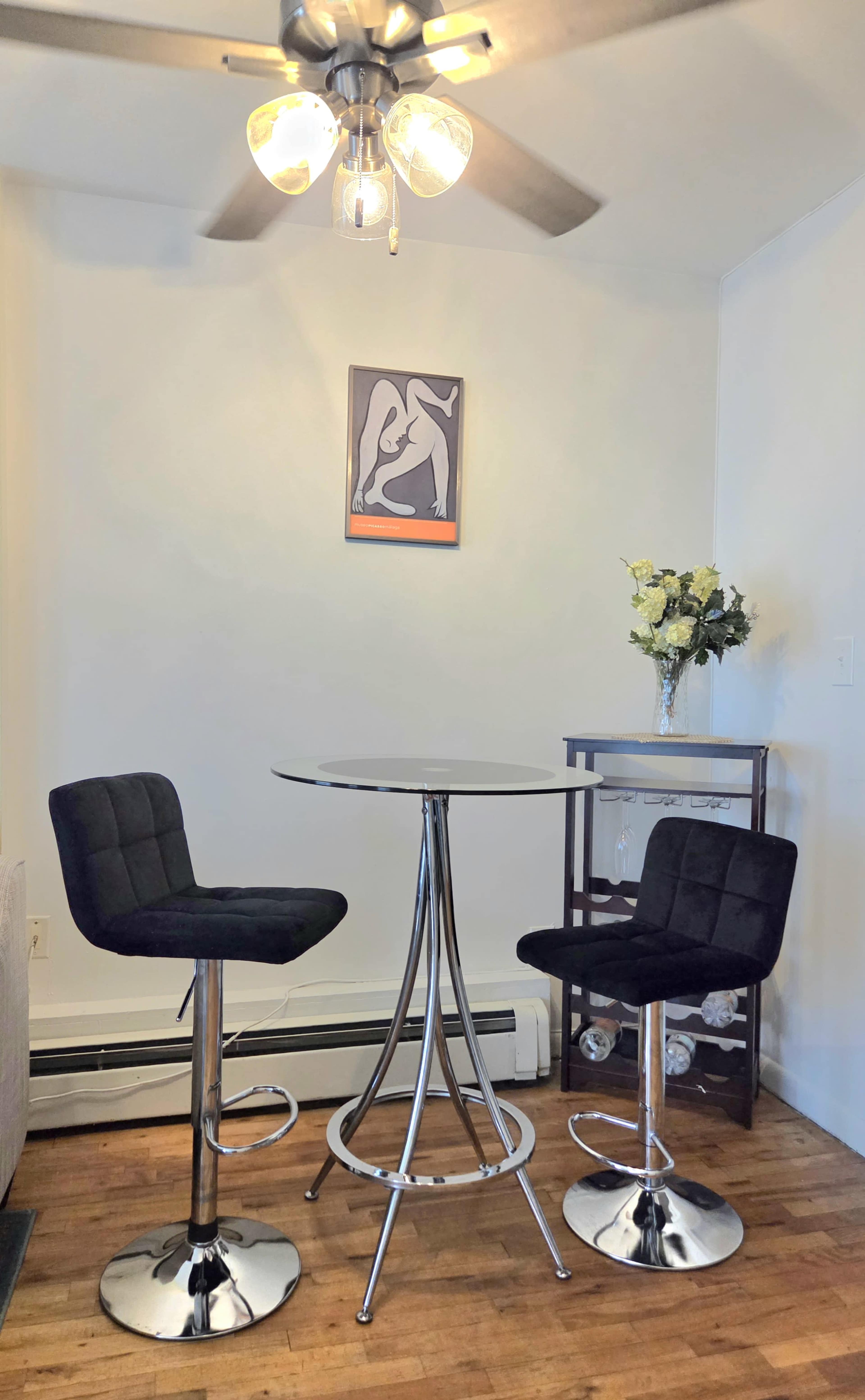 A modern dining area features a round table with two black bar stools under a ceiling fan, accompanied by a wall art piece and a vase of flowers on a nearby cart.