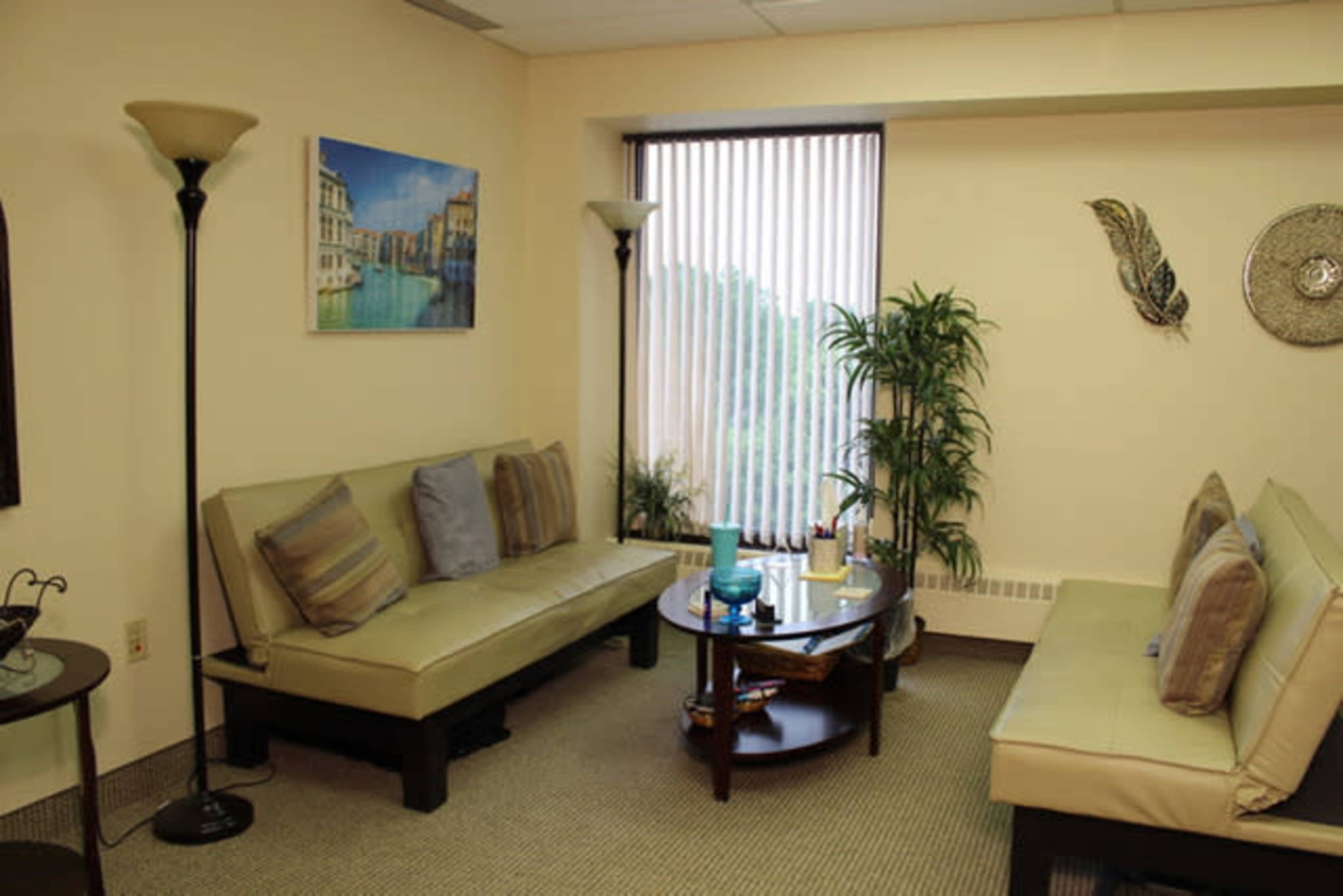 A waiting room with two green sofas, a coffee table, potted plants, and wall art near a large window.