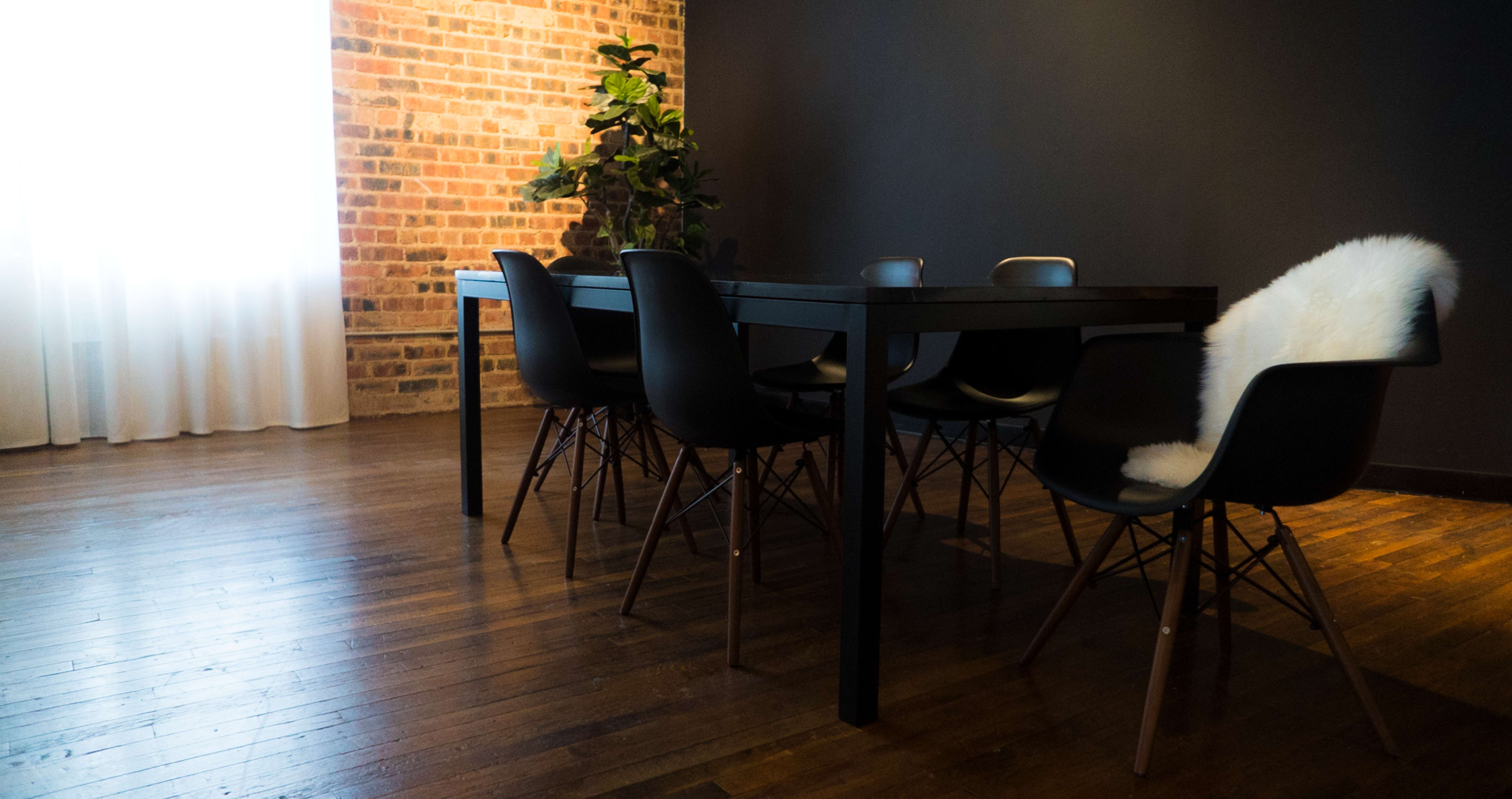 A wooden dining table with black chairs sits in a room featuring exposed brick walls and a large window partially covered by white curtains.
