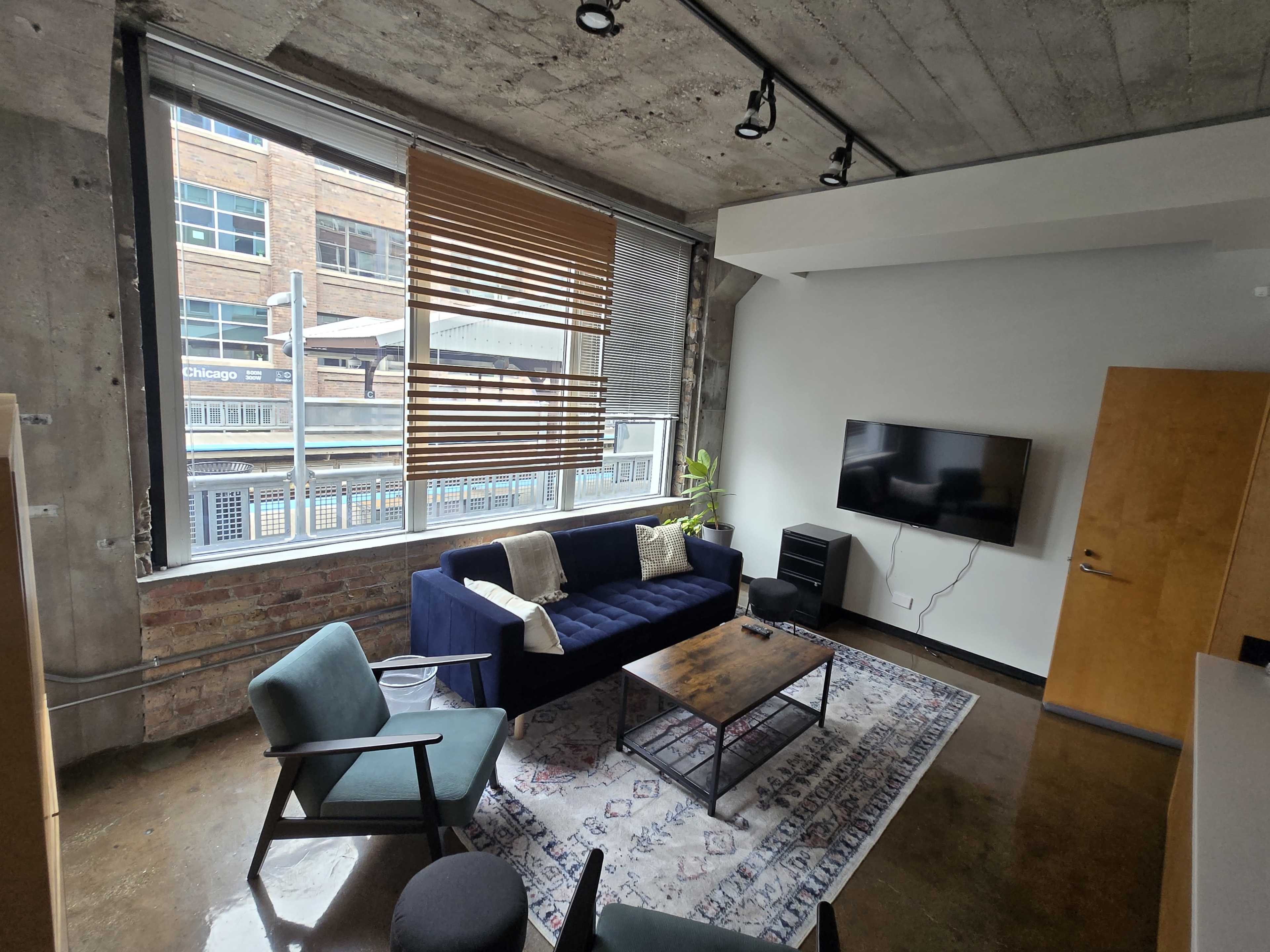 The image shows a modern living room with a blue sofa, a coffee table, a television mounted on the wall, and large windows with blinds allowing natural light in.