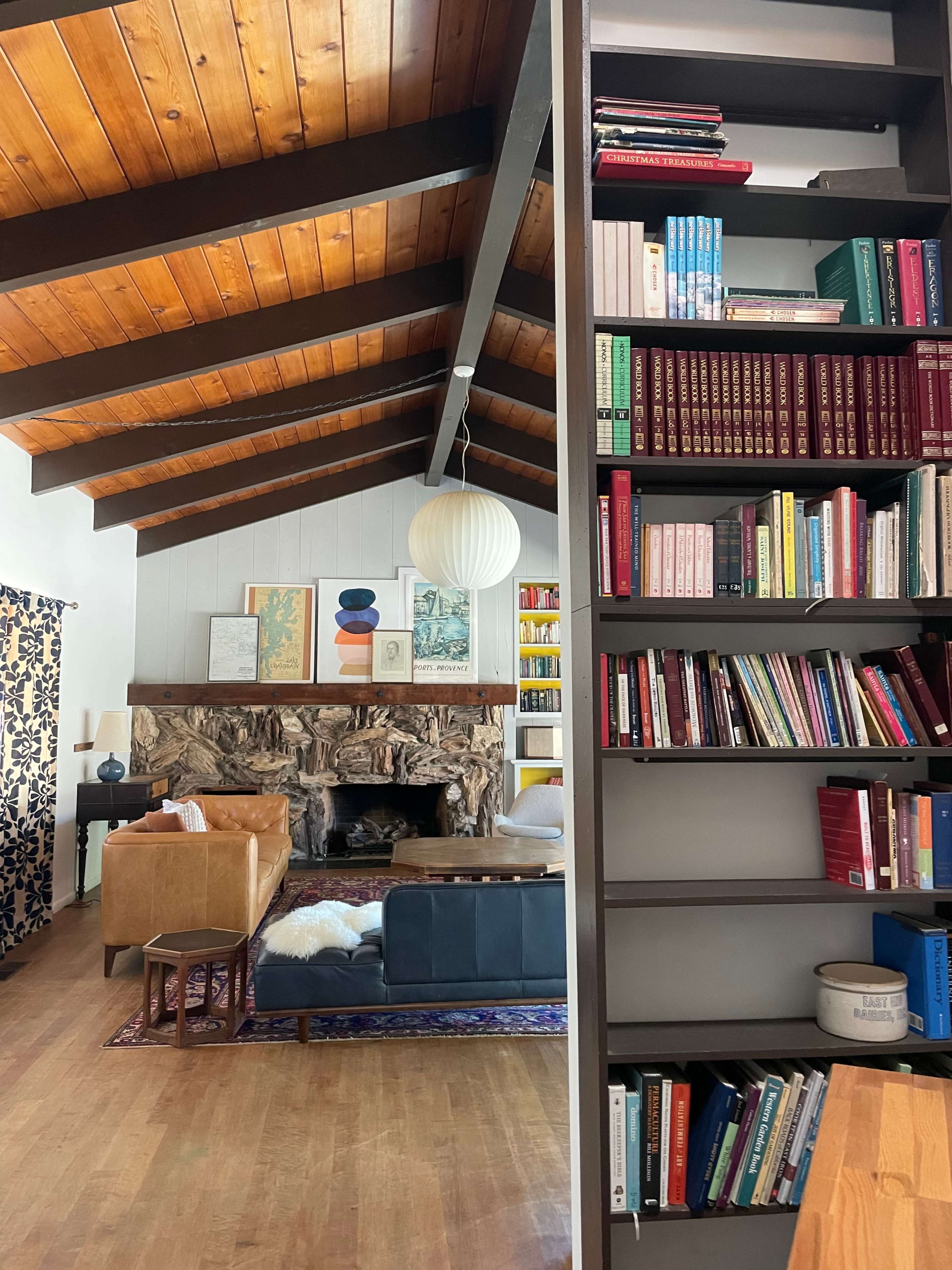 A living room with a high, wooden-beamed ceiling, a stone fireplace, and bookshelves filled with various books.
