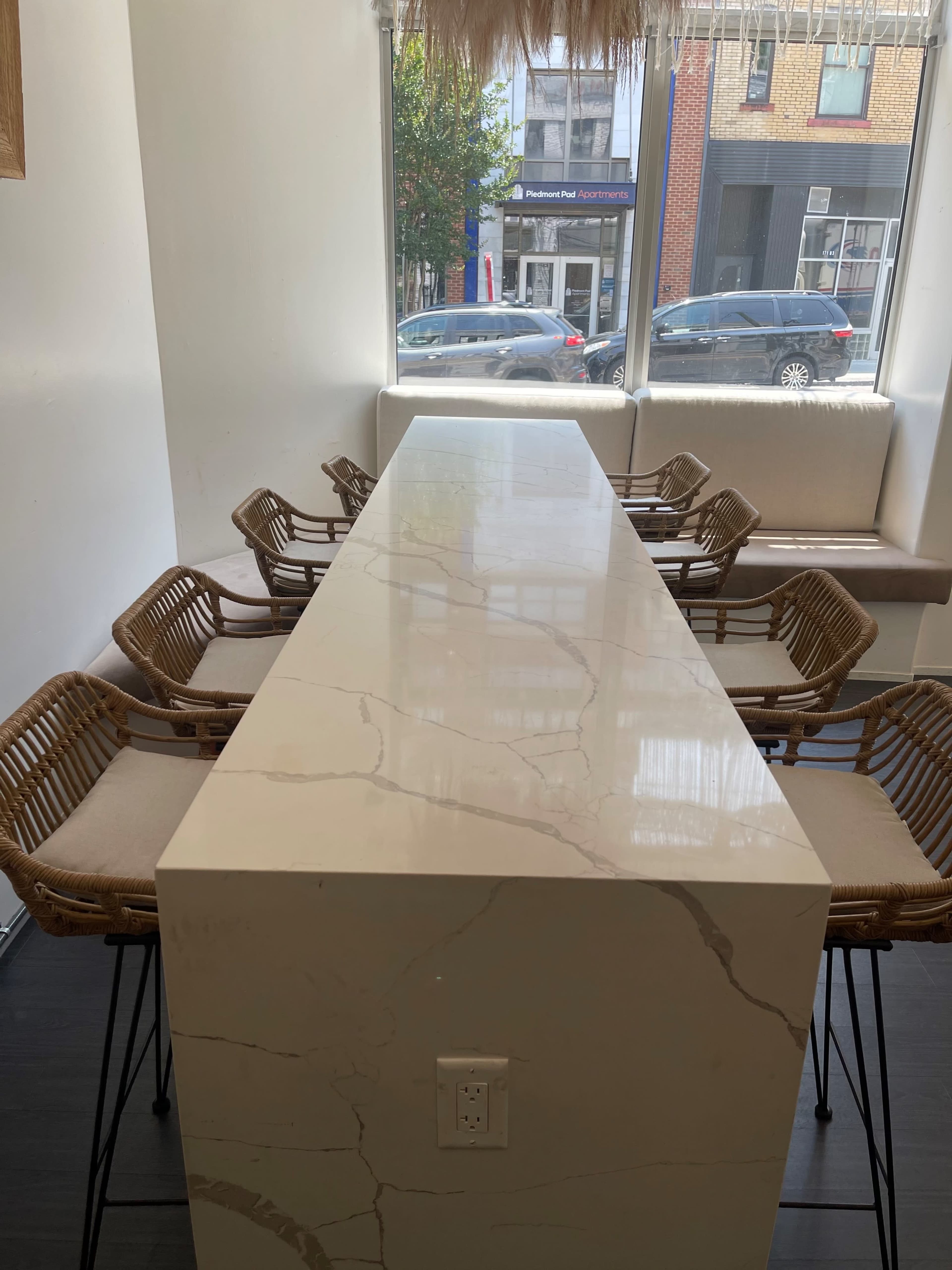 A long, white marble table with rattan chairs is positioned beside a large window in a cafe.