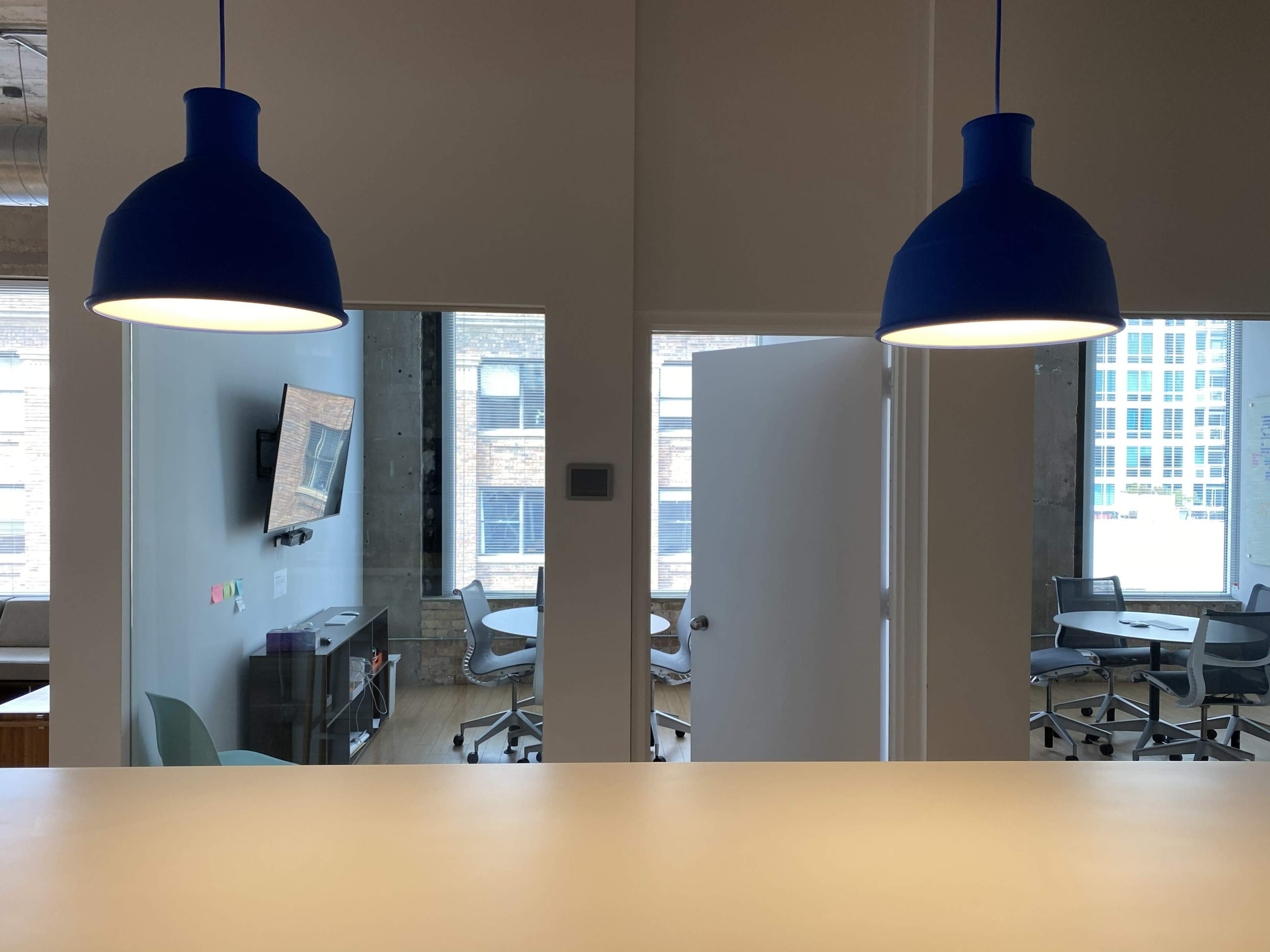 The image shows an office space with blue pendant lights, a visible doorway leading to another room, and modern furniture arranged for meetings.