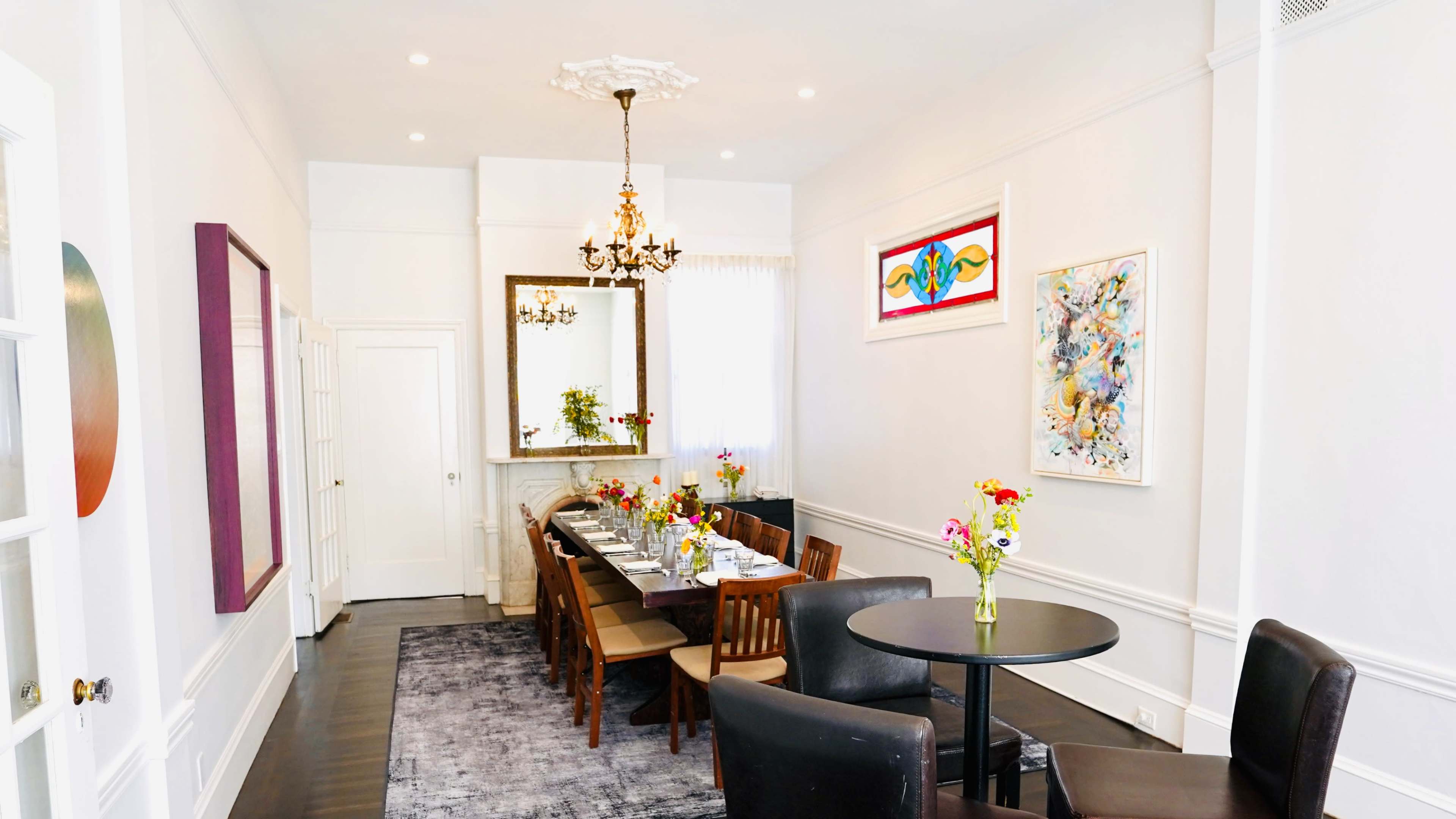 A well-decorated dining room features a long table set for a meal, surrounded by chairs, with colorful floral arrangements and artwork on the walls.