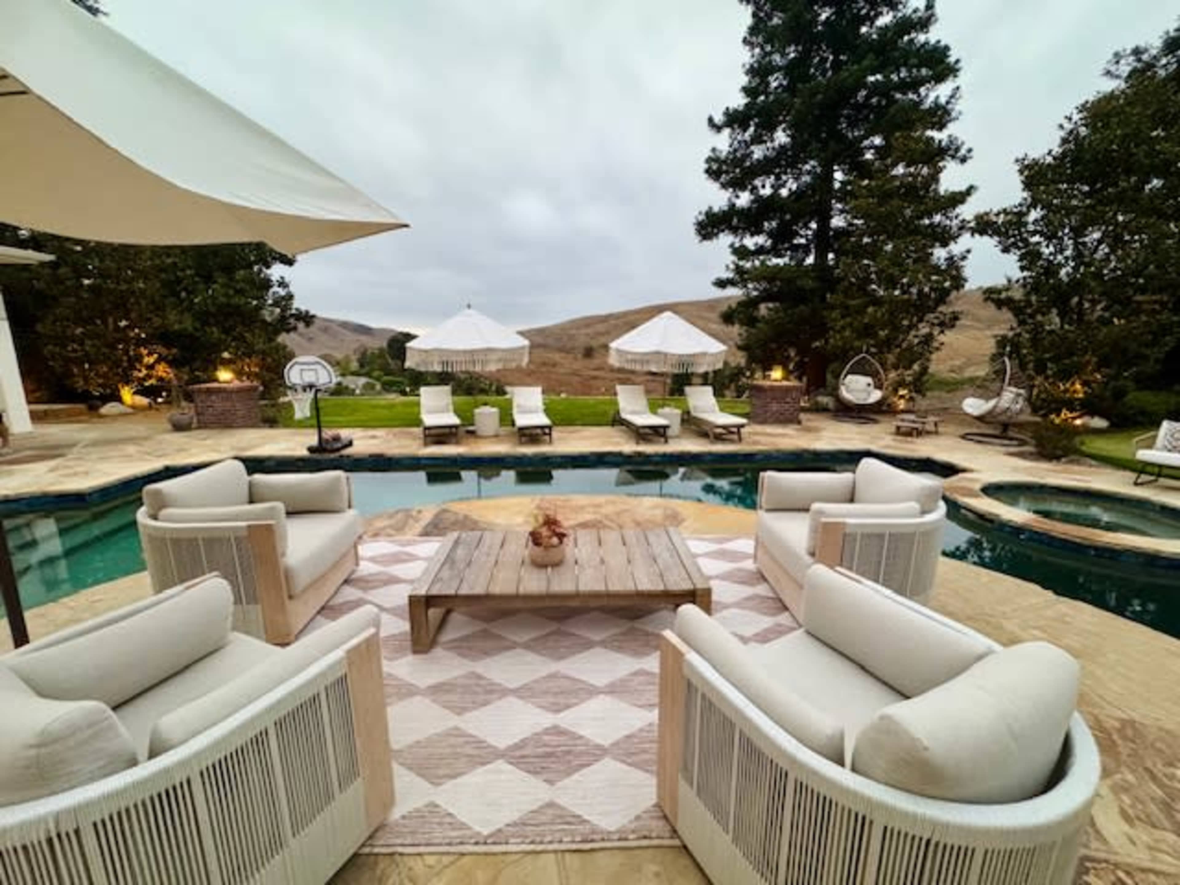 The image shows a cozy outdoor seating area with white chairs arranged around a wooden table, overlooking a pool and lounging chairs under shaded umbrellas.