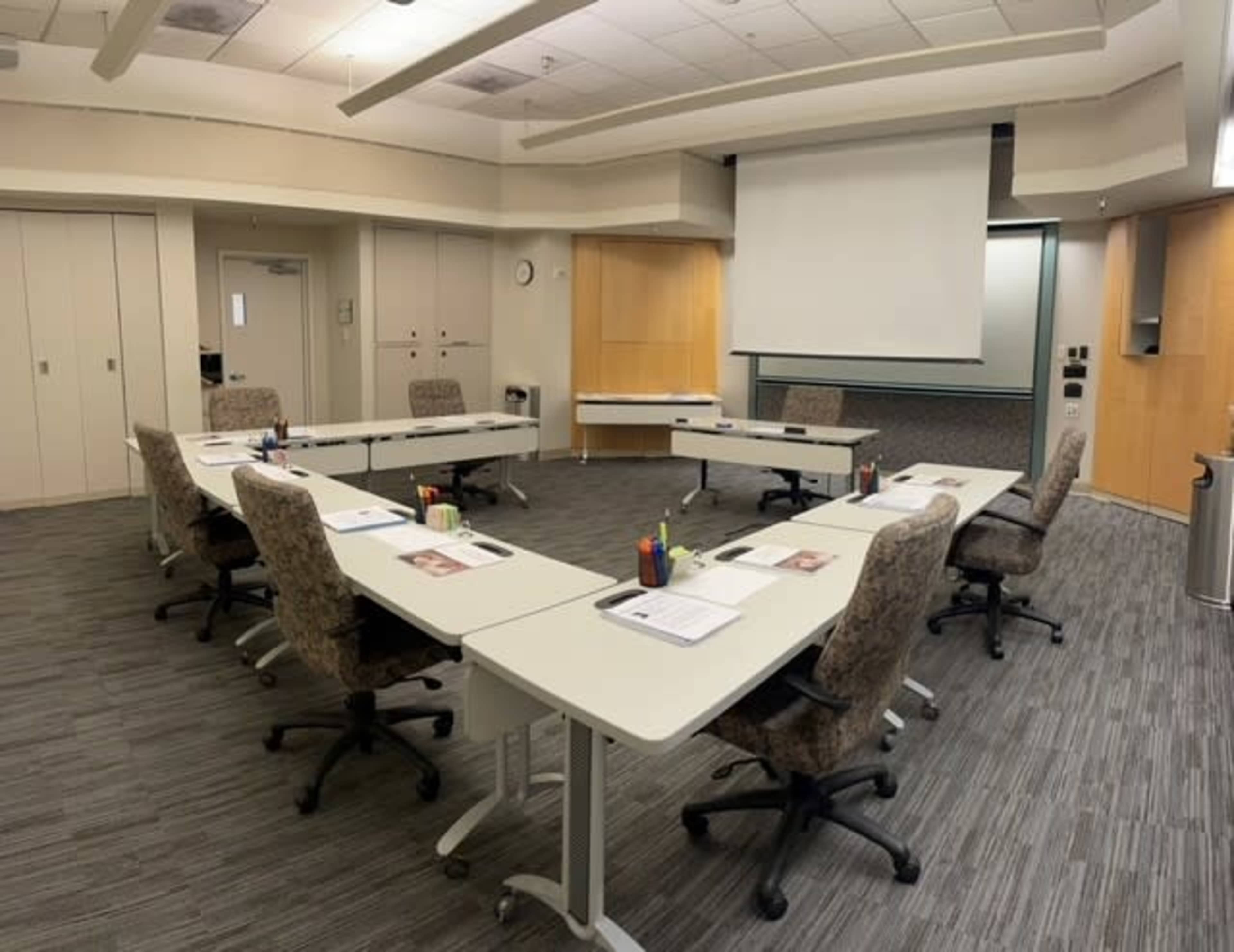 A modern conference room is arranged with several tables in a U-shape, each equipped with chairs, stationery, and a projector screen at the front.