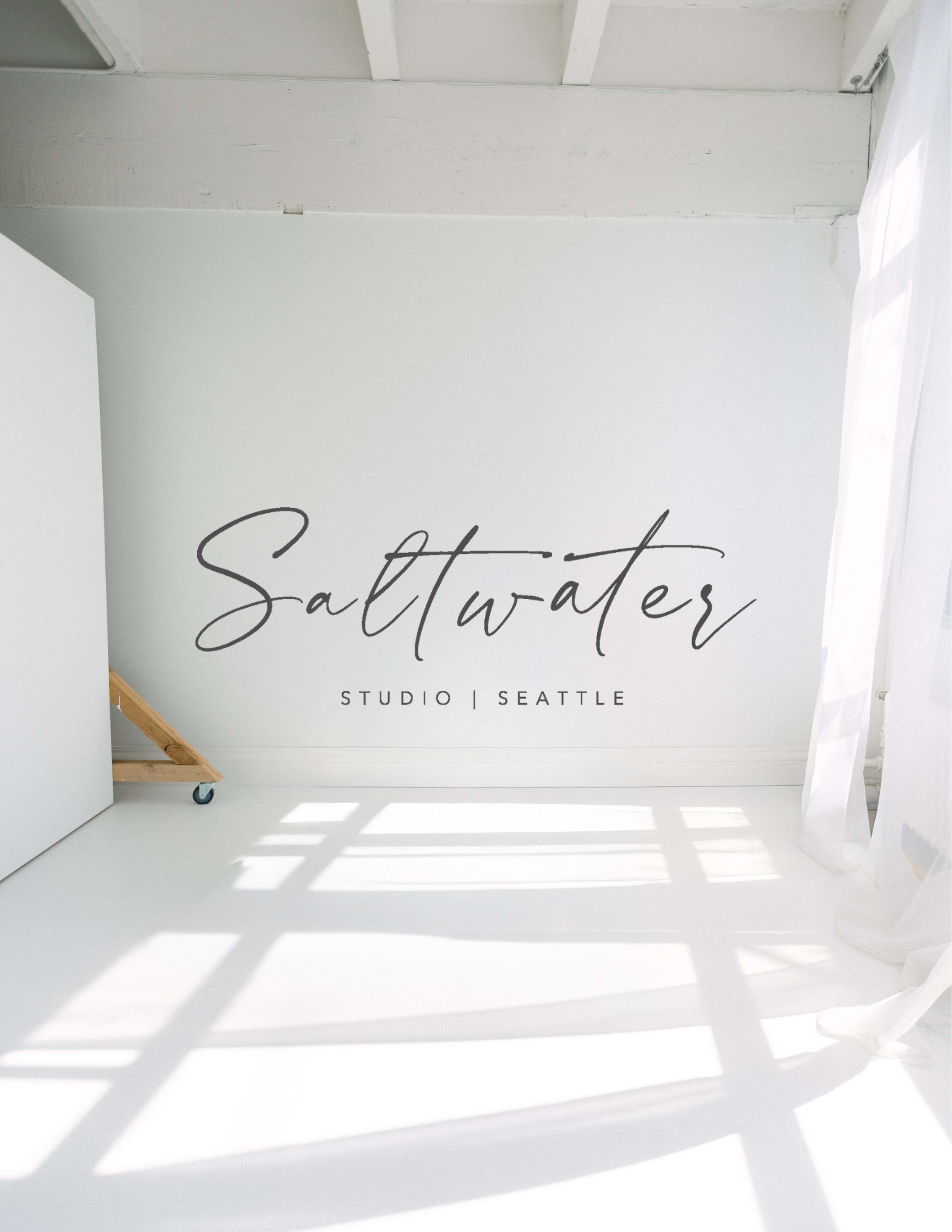 The image shows a bright studio space with a white wall featuring the text "Saltwater Studio | Seattle" and sunlight casting shadows on the floor.