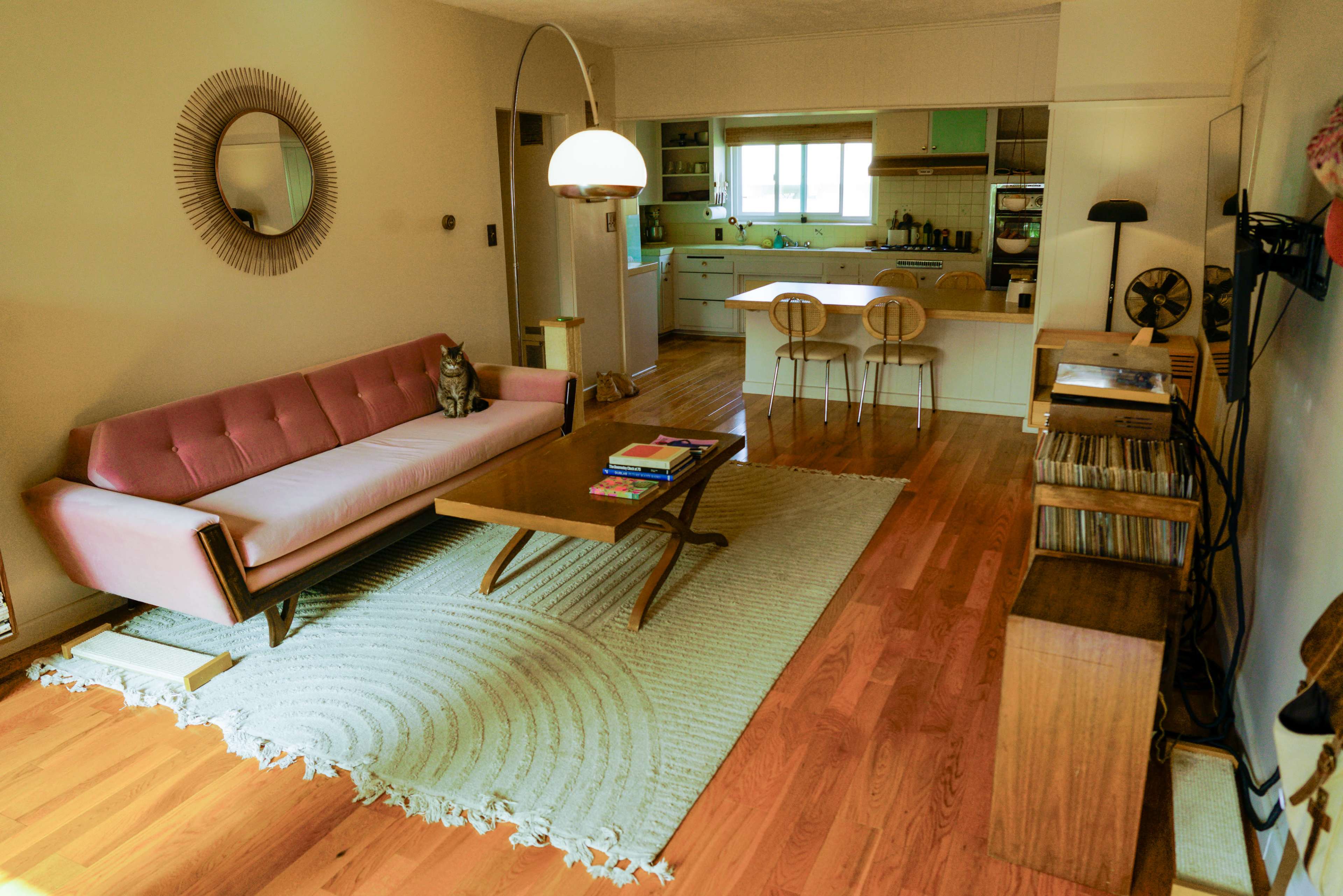 The image shows a living room with a pink sofa, a wooden coffee table, a rug, and an open kitchen in the background.