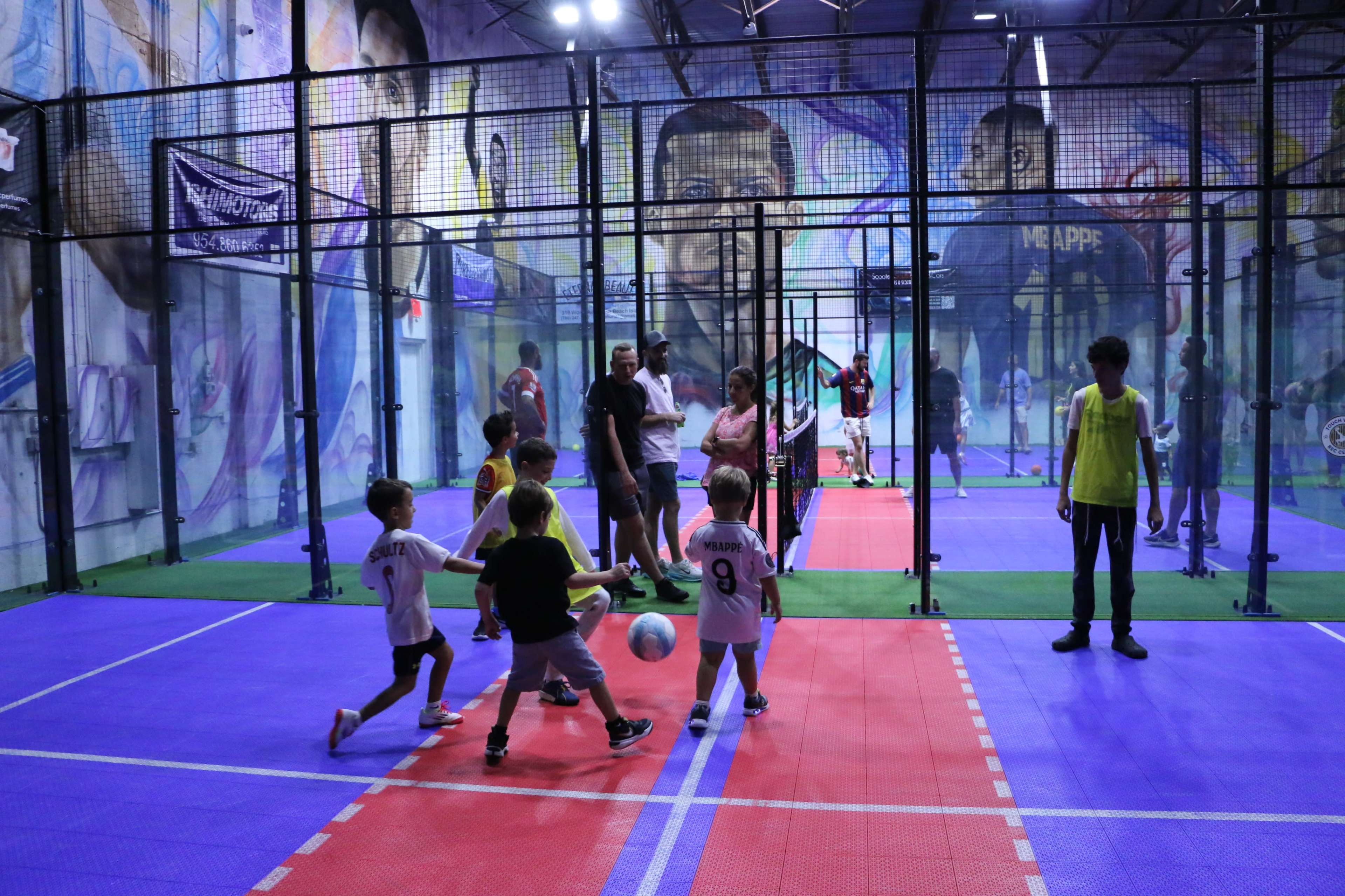 A group of children is playing a small-sided soccer game indoors on a colorful court, surrounded by murals depicting athletes.