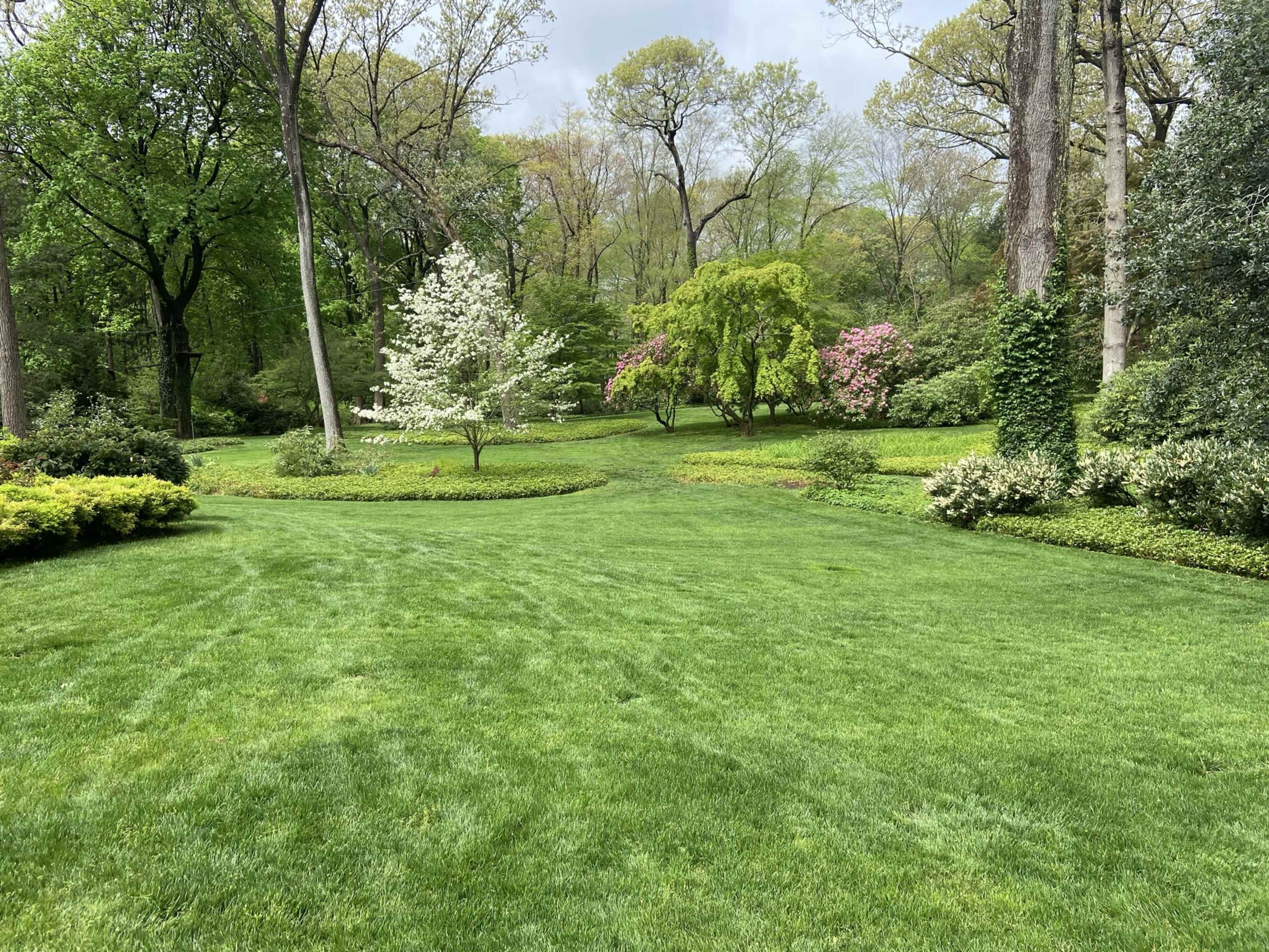 The image shows a manicured garden with lush green grass, several trees, and blooming flowers amidst a well-defined landscape.