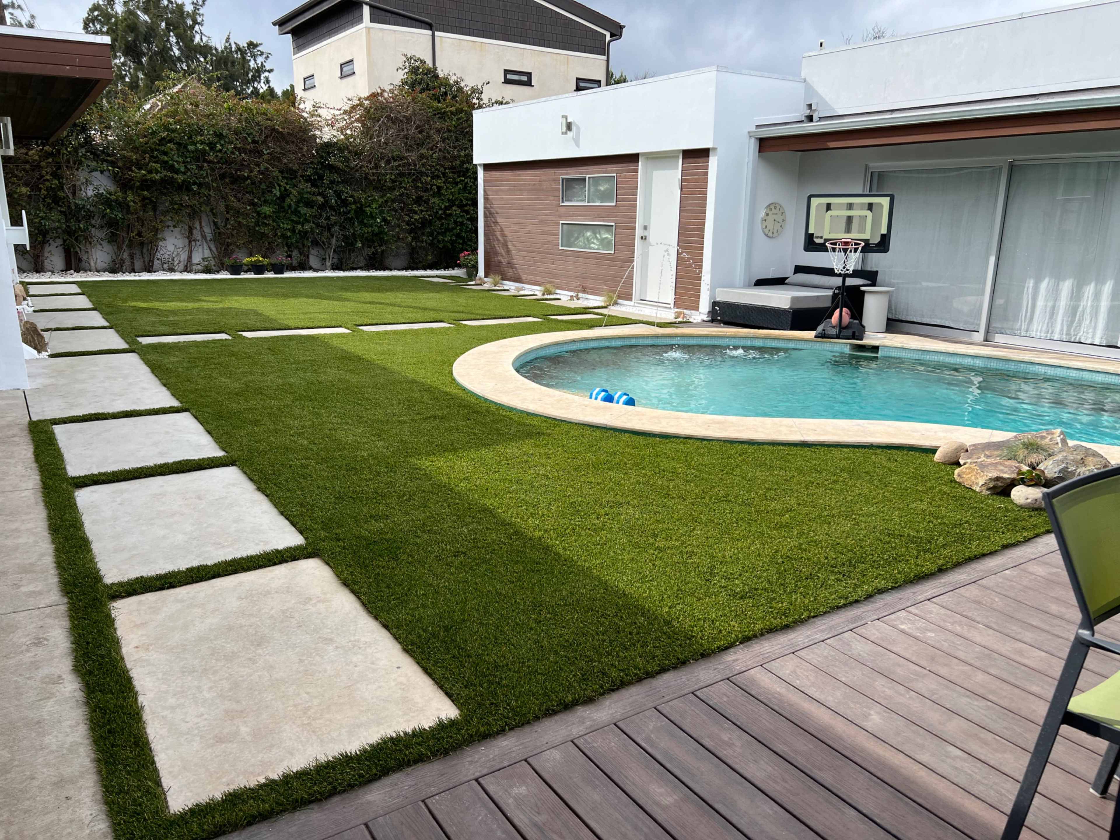 A backyard with a swimming pool, artificial grass, stone pathways, and a basketball hoop mounted near the pool area.