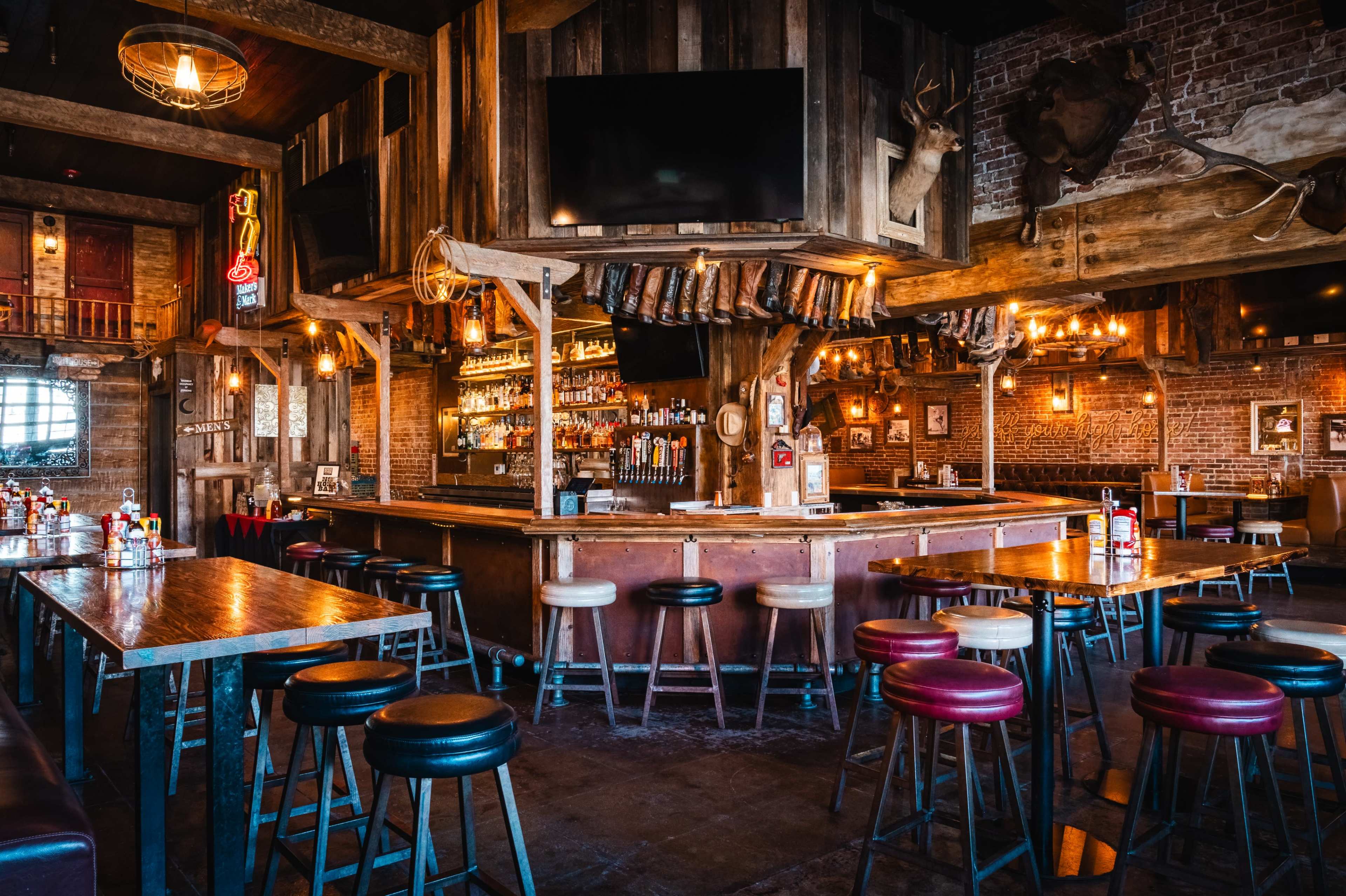 The image shows a rustic bar interior with wooden furnishings, a central bar counter, and shelves lined with various bottles.