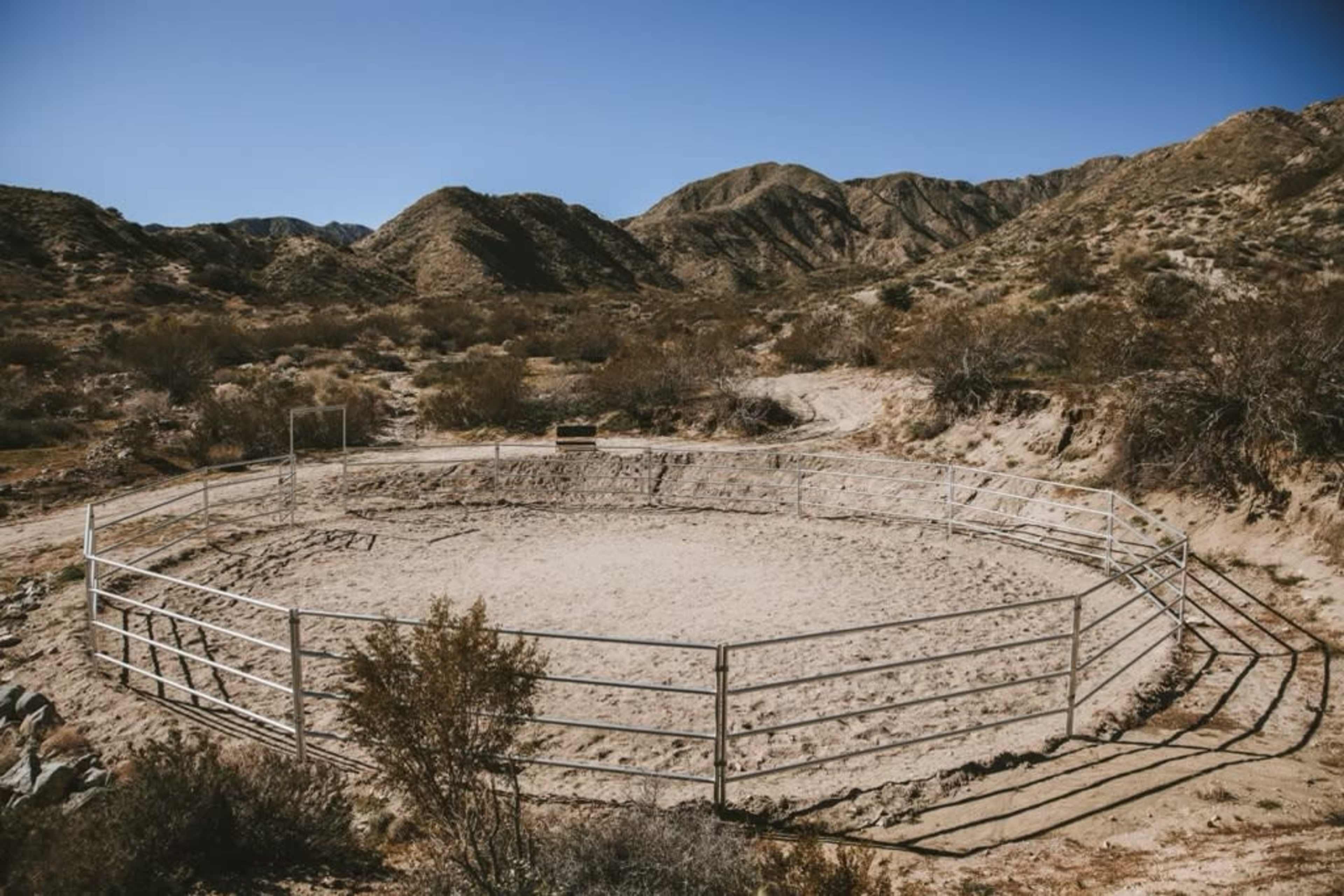 A circular dirt arena enclosed by a metal fence is situated among dry, hilly terrain.
