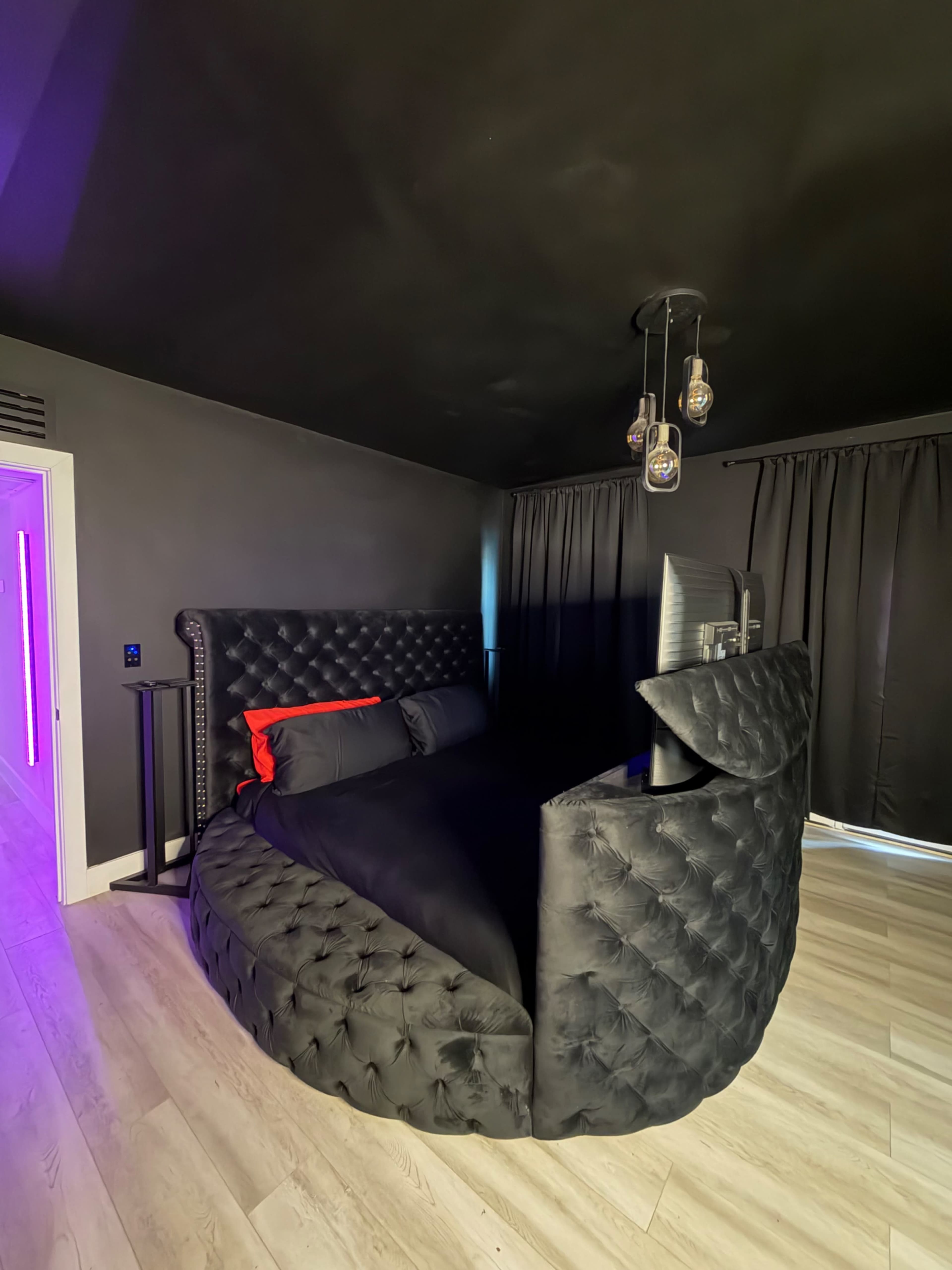 The image shows a circular black bed with tufted upholstery and a headboard, set against a dark room with black curtains and modern pendant lighting.