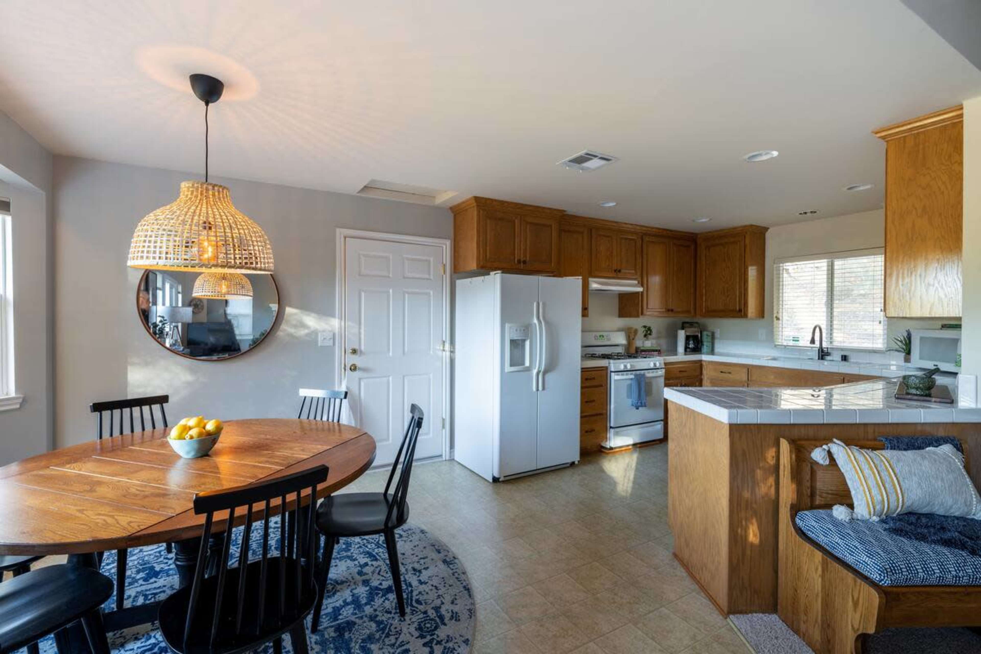 A bright kitchen features wooden cabinets, a round dining table with black chairs, and a large refrigerator next to a window.