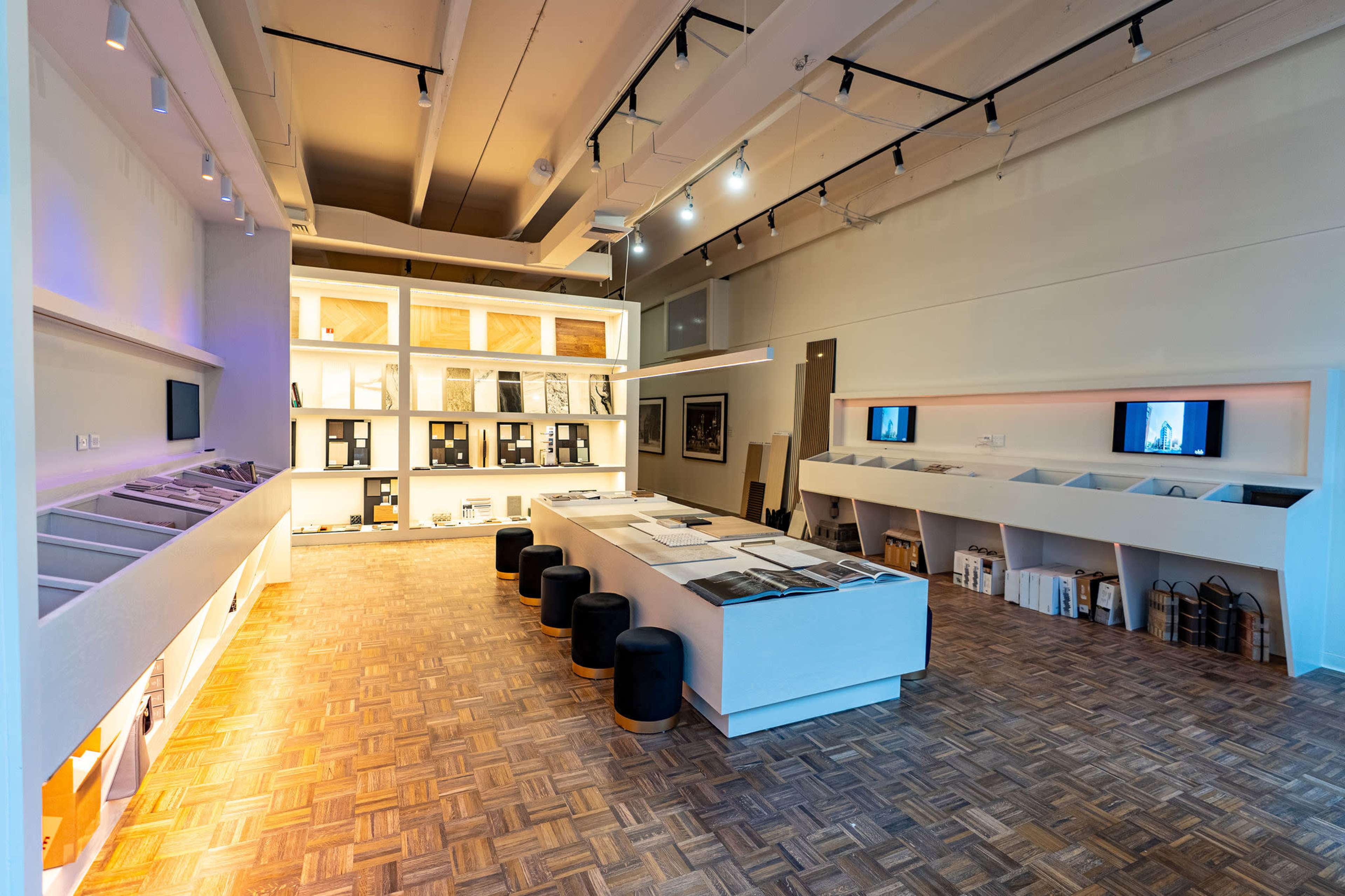 The image shows a modern exhibition space with white walls, a wooden floor, and various display areas featuring architectural materials and screens.