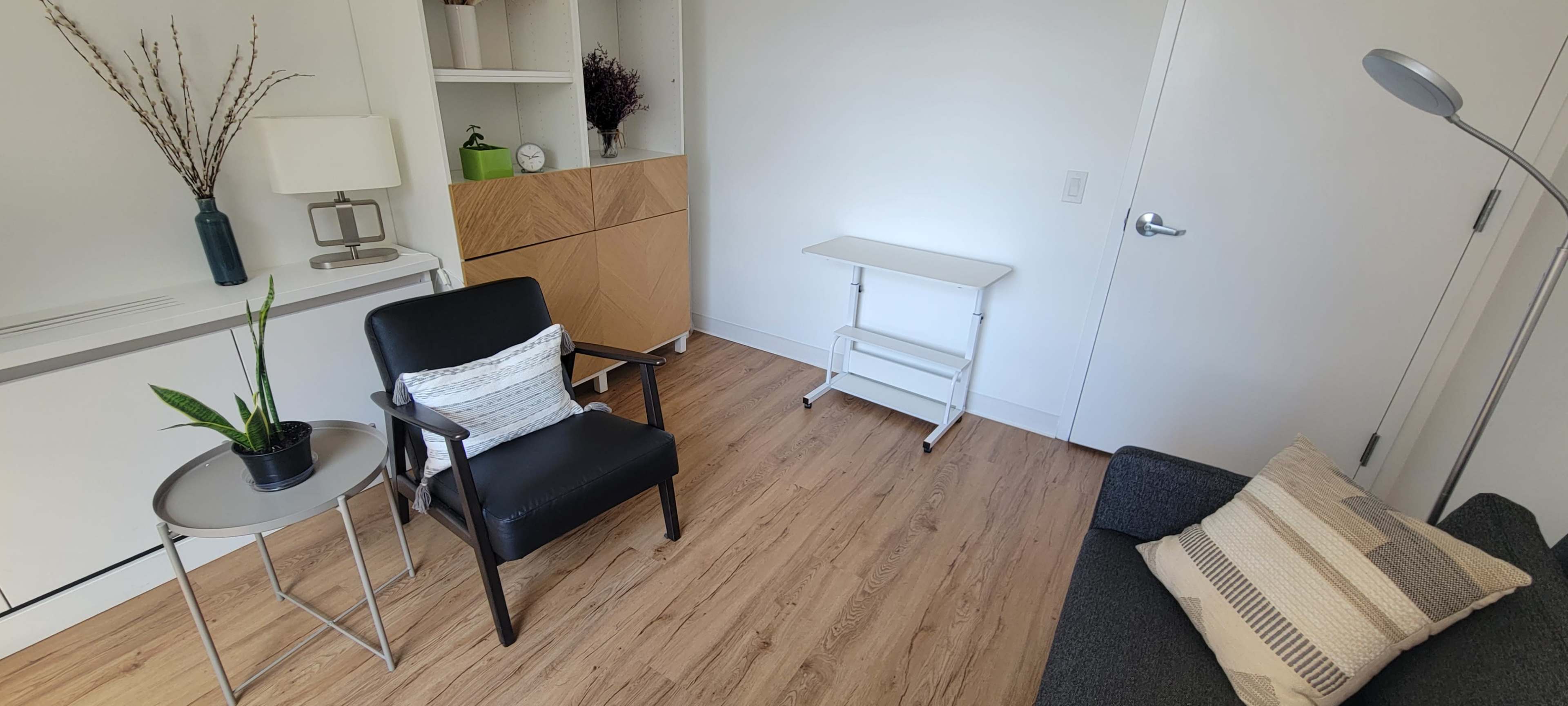A neat room featuring a black chair, a small round table, a white folding table, and a gray sofa against light-colored walls and wooden flooring.