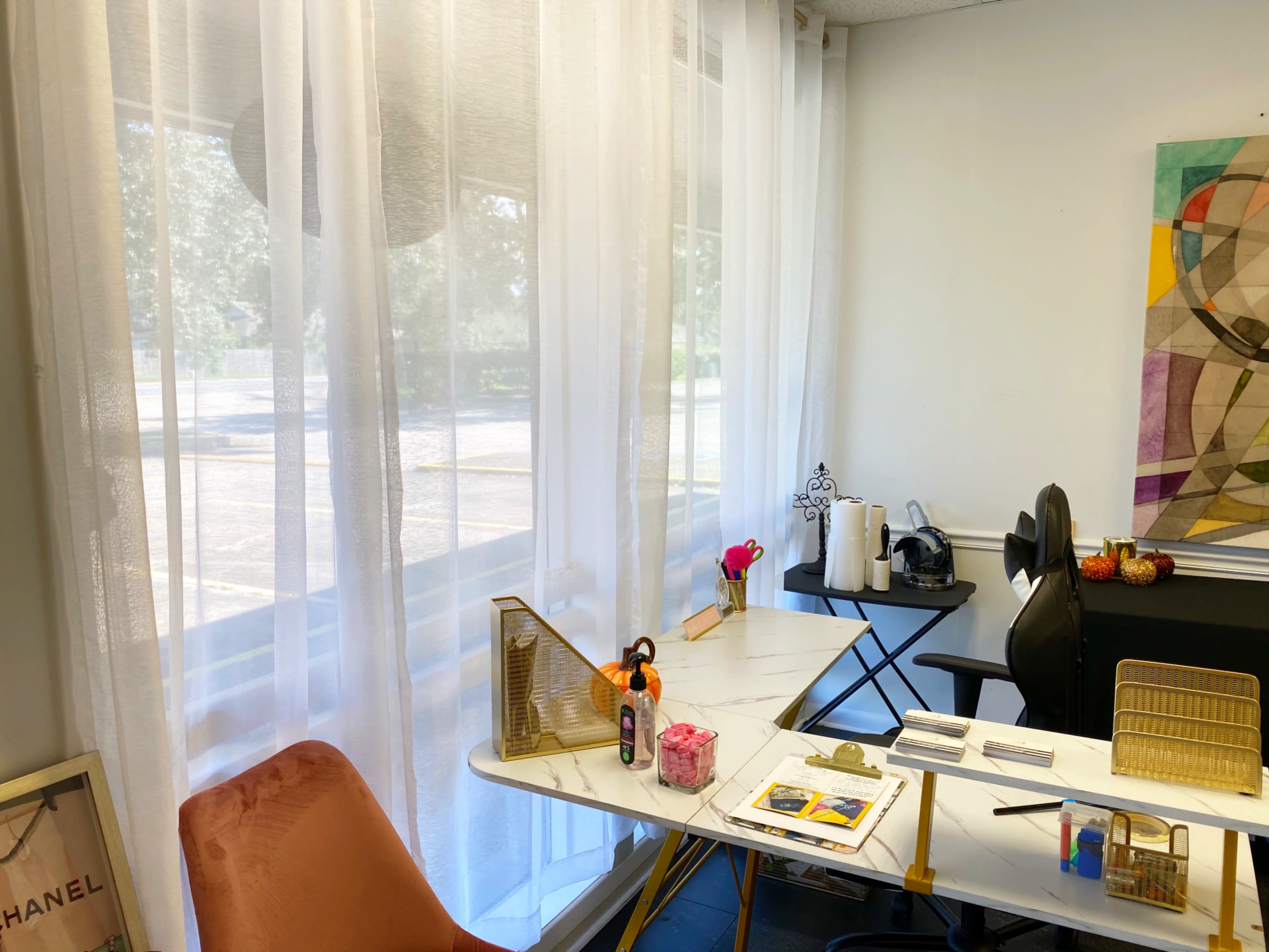 A bright room features a desk with art supplies, a chair, and sheer curtains allowing natural light to filter through.