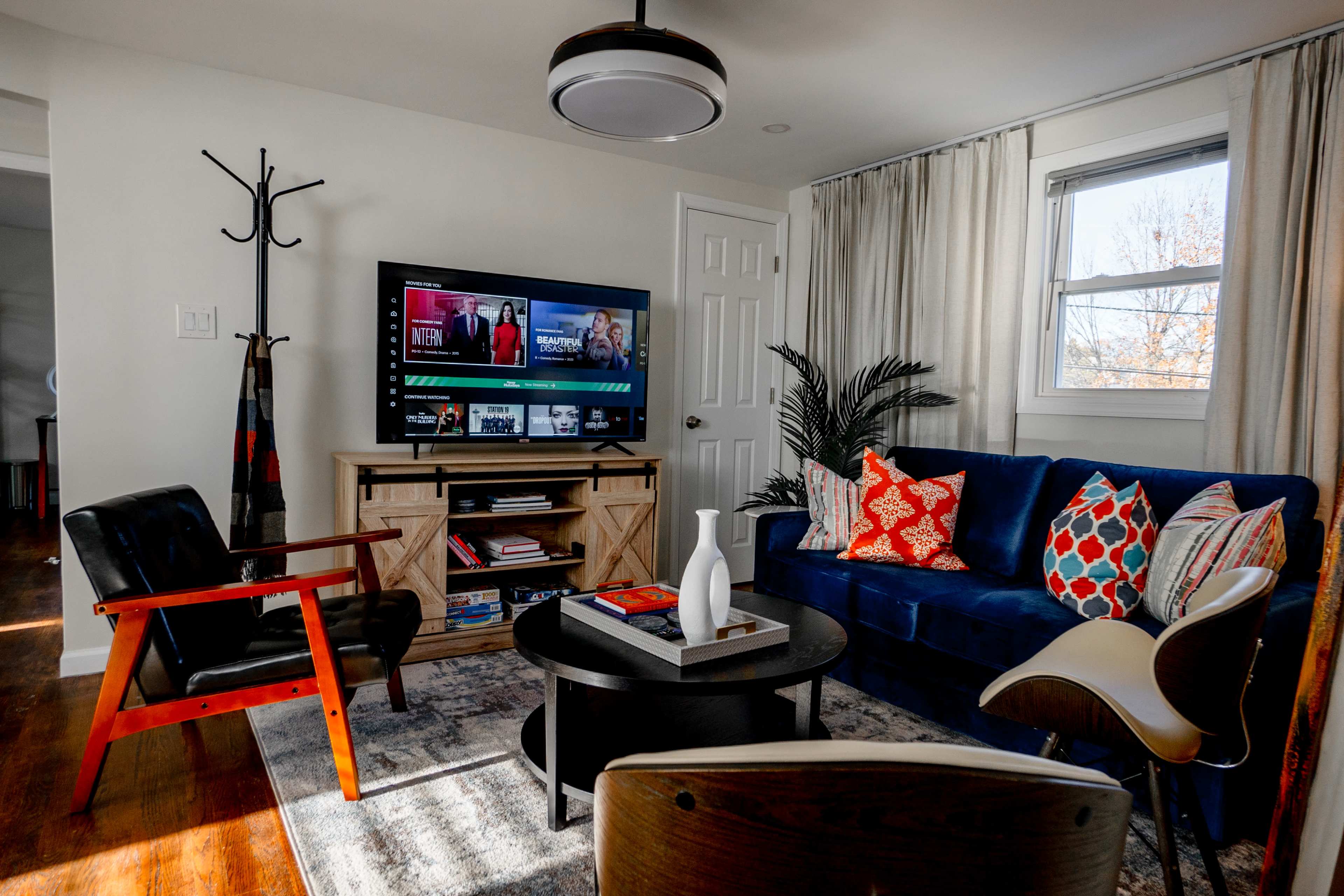 A living room with a blue sofa, a round coffee table, a wooden entertainment center with a television, and various decorative pillows on the sofa.