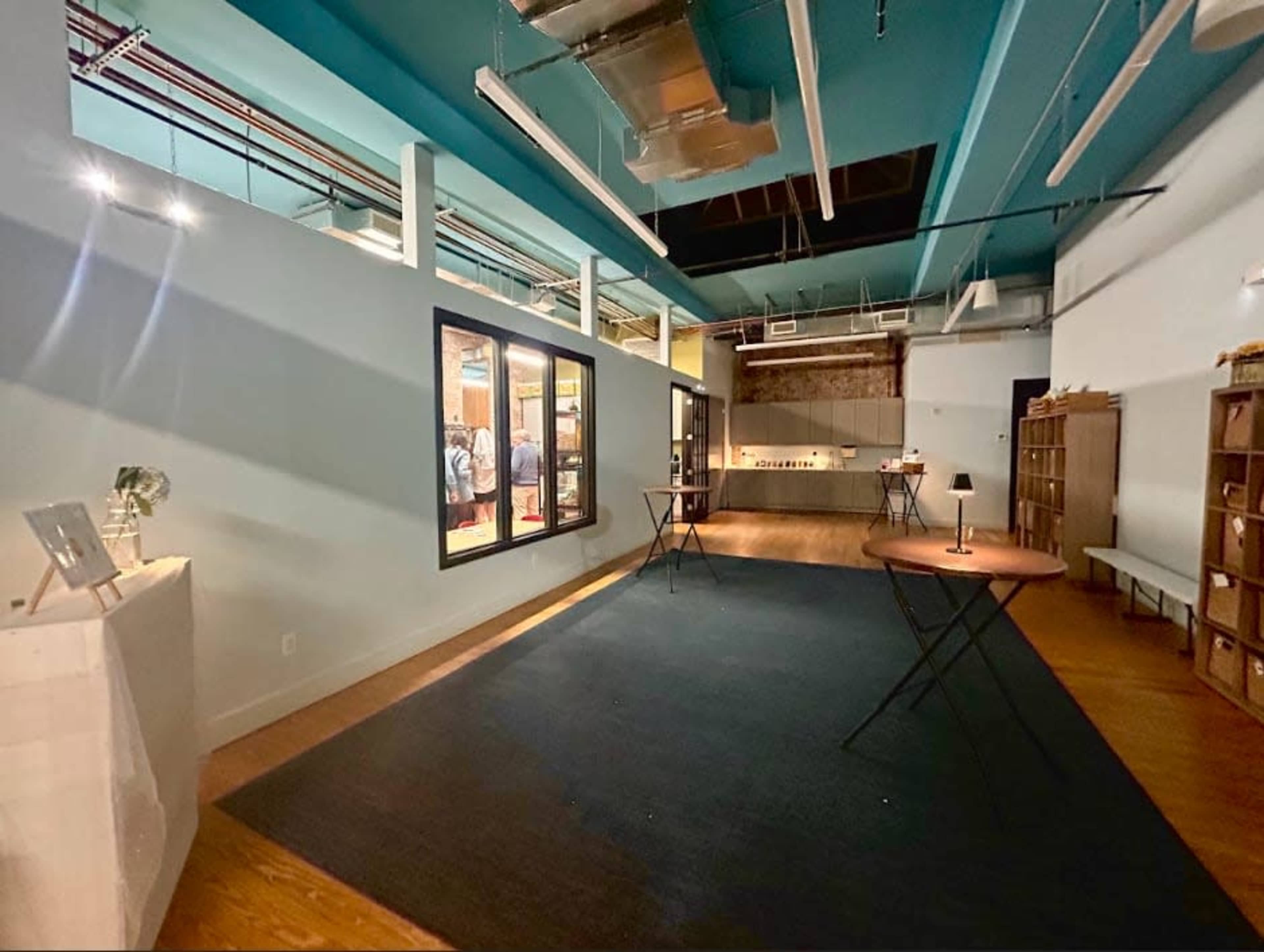 The image shows a spacious, modern interior with high ceilings, wooden floors, and several tables arranged in an open area, along with a shelf against one wall.