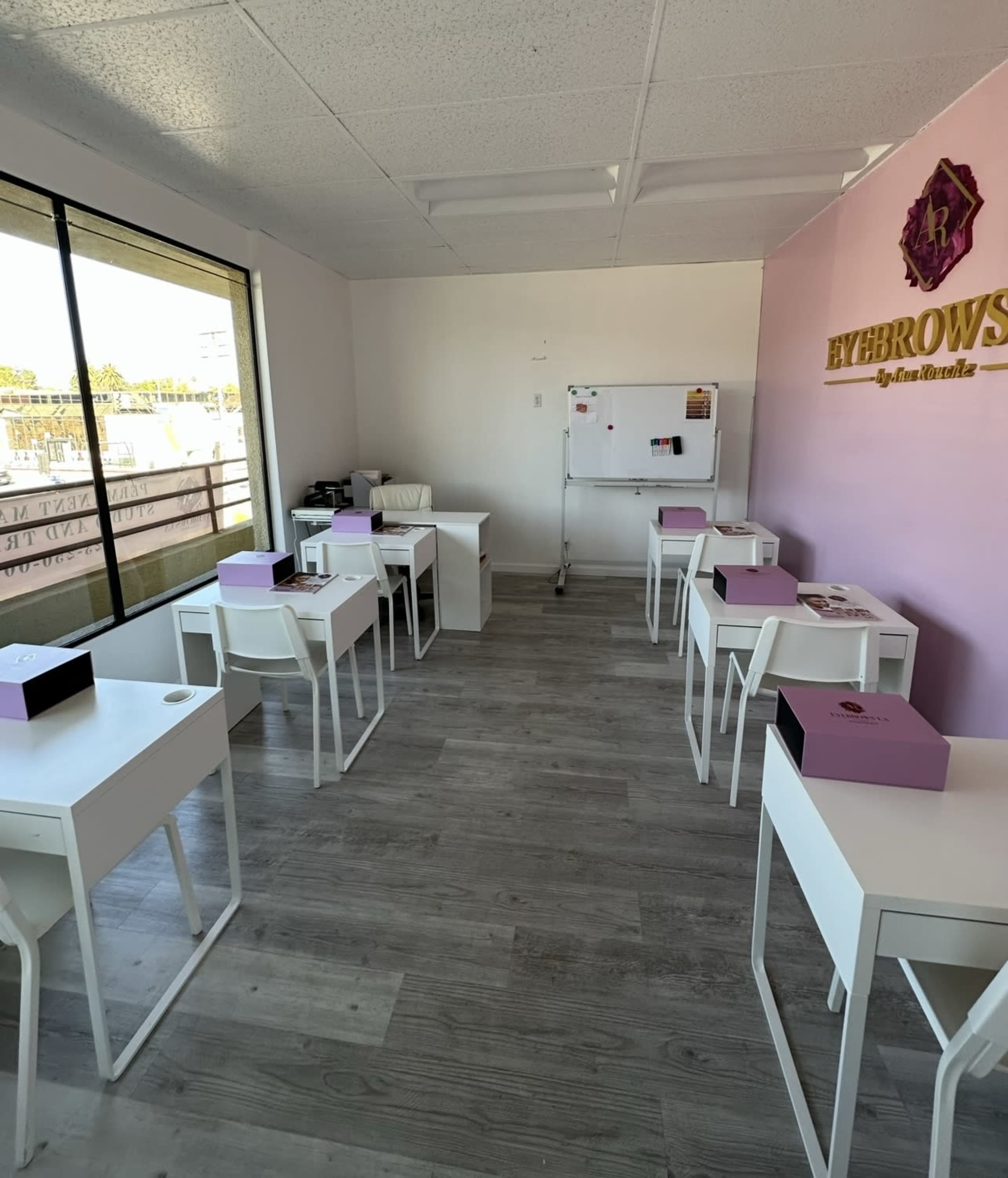 The image shows a bright, organized room featuring several white desks arranged in a row, with a whiteboard and a pink wall displaying the word "EYEBROWS."